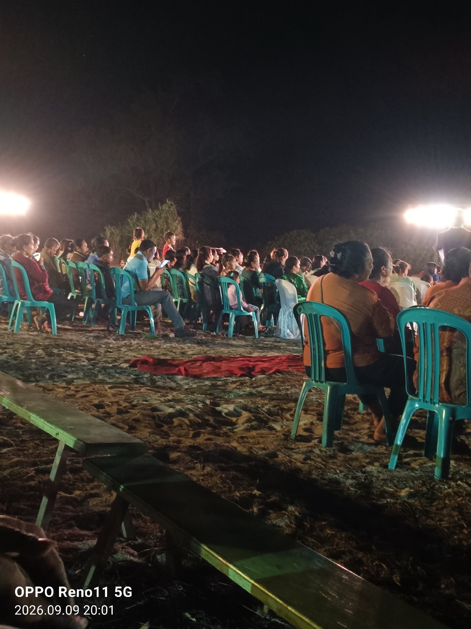 The image depicts an outdoor nighttime scene, likely a gathering or performance, on a sandy surface. A crowd of people are seated on teal-colored plastic chairs, facing towards the right of the frame where bright lights illuminate the area. The lighting suggests a stage or focal point is present, though not directly visible in detail. Some individuals are looking at their mobile phones. In the foreground, a long wooden bench with metal legs is positioned diagonally, partially obscuring the view of some chairs and the sandy ground. The environment appears somewhat rustic, with sparse vegetation visible in the background and sandy, uneven terrain underfoot. The timestamp "OPPO Reno11 5G 2026.09.09 20:01" is present in the bottom left corner, indicating the image was taken with a specific phone model on September 9, 2026, at 8:01 PM. The location is stated as Otan, Indonesia.