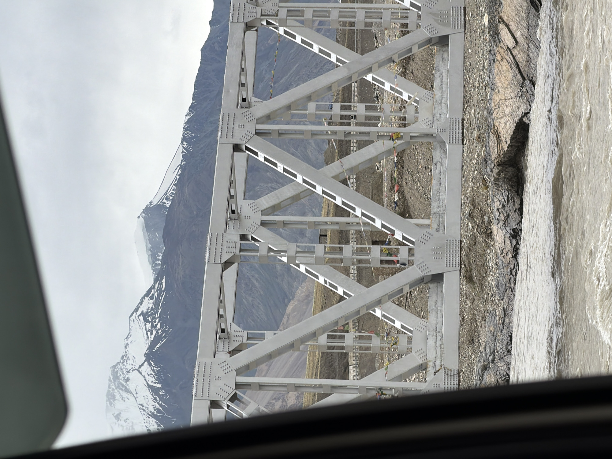 The image shows a white truss bridge spanning a rocky riverbed in Zangla, India. Snow-capped mountains form the backdrop, with a cloudy sky overhead. The bridge is constructed from metal beams and rivets, with diagonal supports creating a geometric pattern. Prayer flags are strung along the bridge, fluttering in the wind. The river below is murky brown and appears to be flowing rapidly. The foreground is dominated by the river and the rocky shore, while the bridge and mountains are visible beyond. The perspective suggests the photo was taken from a vehicle.