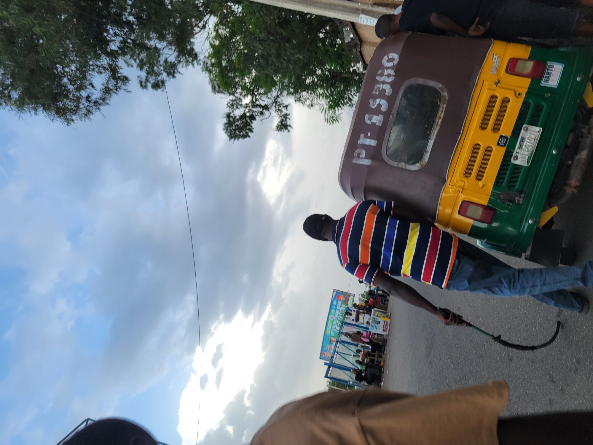 The image captures a street scene in Jos, Nigeria, with a focus on a three-wheeled vehicle, commonly known as a "keke," and several individuals. The keke, predominantly brown and yellow with green accents, is positioned at an angle. Its registration plate is visible, displaying the characters "JUN-579YC." The side of the keke bears white lettering that appears to read "PT-15360."
A man in a striped shirt (blue, orange, white, and yellow) and blue jeans is seen holding a curved, dark object in his right hand, which he is dragging on the asphalt road behind him. His posture suggests he is walking away from the viewer and towards the keke. Another man, wearing a black t-shirt, is partially visible on the right side of the image, leaning towards the back of the keke. The background reveals a bustling area with a billboard displaying colorful imagery and text, including portraits of individuals. Several other people are seated or standing near what appears to be a roadside stall or gathering point. Trees and utility lines are also discernible in the broader environment, under a cloudy sky. The ground is paved with asphalt, indicating a road or a marketplace.