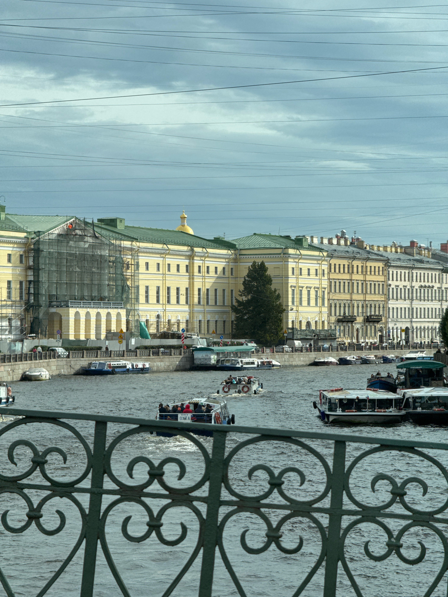 This is an eye-level, medium shot of a canal in Saint Petersburg, Russia, with a building facade and a decorative railing in the foreground. The canal is filled with several boats, including tour boats and smaller vessels. On the left bank of the canal, there is a large, yellow building undergoing renovation, covered in green scaffolding and netting. Further down the canal, more yellow and white buildings with green roofs line the bank. A dark green, ornate railing runs across the bottom of the frame, partially obscuring the view of the canal and boats. The sky is overcast with a few visible clouds. The overall scene depicts a city canal with architectural elements and active water traffic under a cloudy sky.
