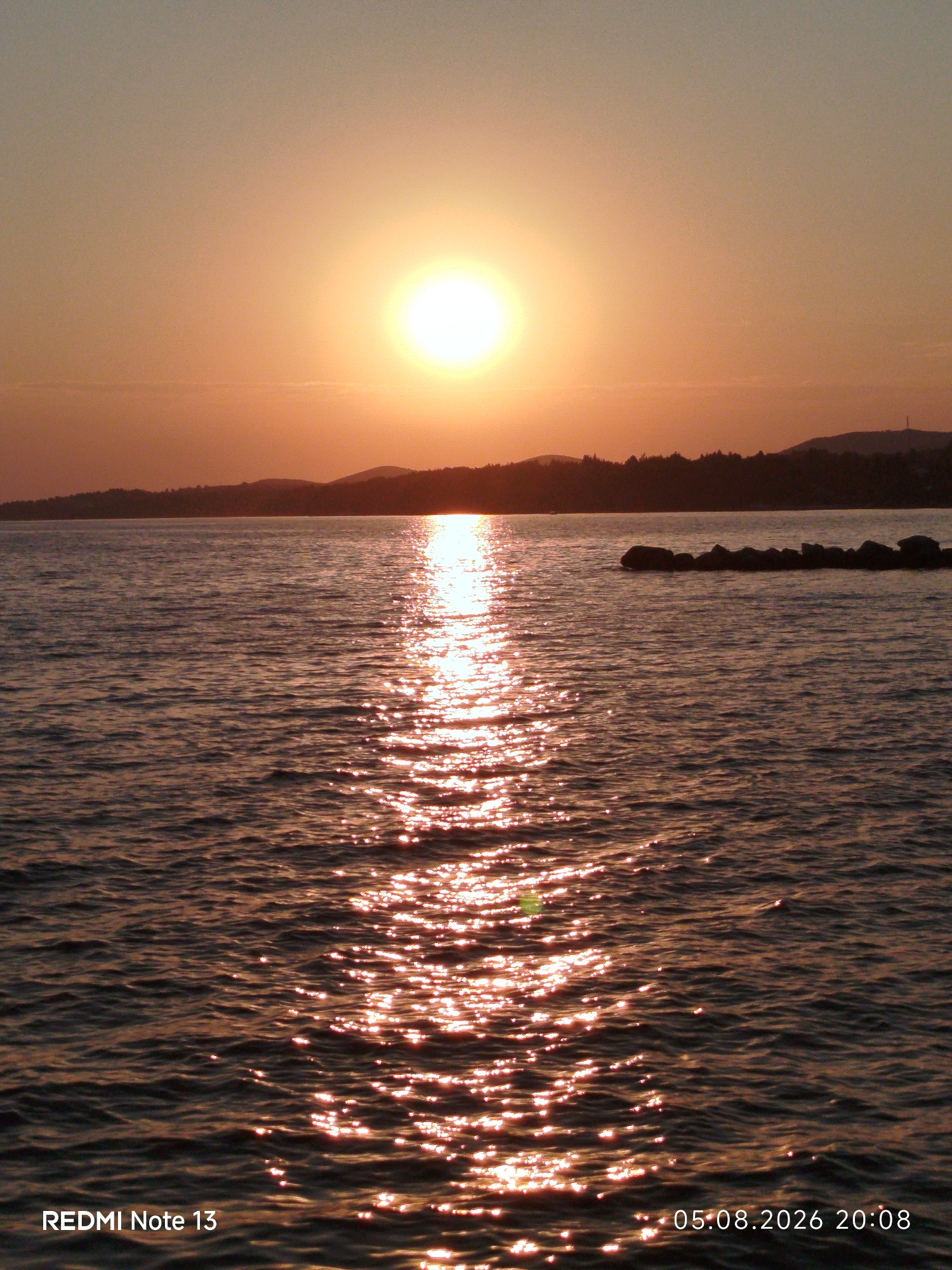 A sunset over the sea is captured in this vertical, full-frame photograph. The sun, a bright, circular orb with a distinct halo, is positioned slightly left of center in the upper portion of the frame, casting a warm, golden glow across the scene. A shimmering reflection of the sun stretches across the water towards the viewer, creating a pathway of light. The sea itself is calm, with gentle ripples disturbing its surface. In the background, a silhouetted coastline with rolling hills and scattered trees forms a dark horizon line against the orange and pale yellow sky. A breakwater, made of dark, irregular rocks, is visible in the mid-ground, breaking the expanse of water. Text overlays at the bottom left and bottom right of the image read "REDMI Note 13" and "05.08.2026 20:08" respectively, indicating the device used and the date and time of the photograph. The overall atmosphere is serene and peaceful, characteristic of a coastal sunset in Sithonia Municipal Unit, Greece.