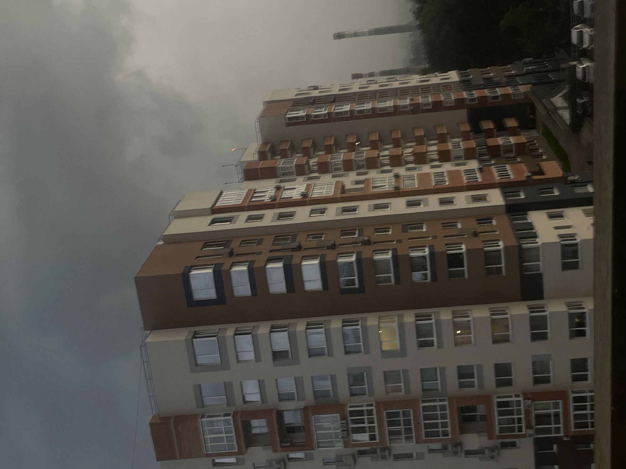 The image captures a towering, multi-story apartment building with a striking architectural design featuring alternating brown and light-colored sections. The building appears to be residential, evidenced by the numerous windows visible on each level. Some windows are open, suggesting the presence of inhabitants. The structure is long and extends out of frame, giving a sense of its scale.
The setting appears to be an urban or suburban environment, typical for apartment complexes. There are no people clearly visible in the image. The building's facade is detailed with balconies, recessed sections, and varying window styles, some of which are large and multi-paned, while others are smaller and more uniform. The overall impression is one of modern construction.
The sky is overcast and a dark, stormy gray, indicating inclement weather, likely rain or impending rain, and contributing to a somber mood. The time of day is difficult to ascertain definitively due to the cloud cover, but the light suggests it is either early morning or late afternoon.
A tall, cylindrical structure, possibly a smokestack or industrial pipe, is visible in the background against the dark sky, along with hints of trees and other indistinct structures. The perspective of the shot is from a slightly elevated position, looking upwards and across at the building.
No visible text is present in the image. The location context of Votkinsk, Russia, helps place this modern residential building within that specific geographical area.