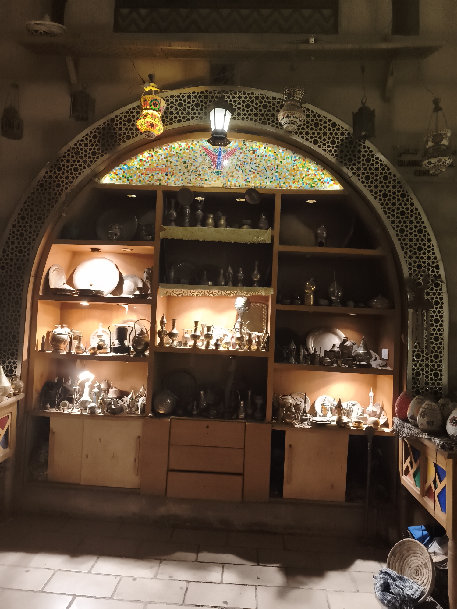 The image depicts an indoor scene, likely a shop or a display area, featuring a large arched wooden shelving unit filled with various decorative items. The shelving unit is illuminated by warm, embedded lights, casting a soft glow on the objects. The items on display include numerous antique-looking metal vessels, such as teapots, pitchers, and decorative containers, as well as plates, bowls, and other intricate metalwork pieces. Some shelves also contain glass items and decorative lamps.
Above the shelving unit, an arched decorative element with a geometric pattern is visible. Suspended from the ceiling are several ornate lanterns, one of which is brightly colored with mosaic glass. To the right of the shelving unit, a sign with Arabic and English text, including "ISLAMIC CENTER," is partially visible. The foreground shows a tiled floor and some woven baskets and textiles. The overall ambiance suggests a place that showcases cultural artifacts, possibly in Sharm El Sheikh, Egypt, given the presence of Arabic script. There are no people visible in the image.