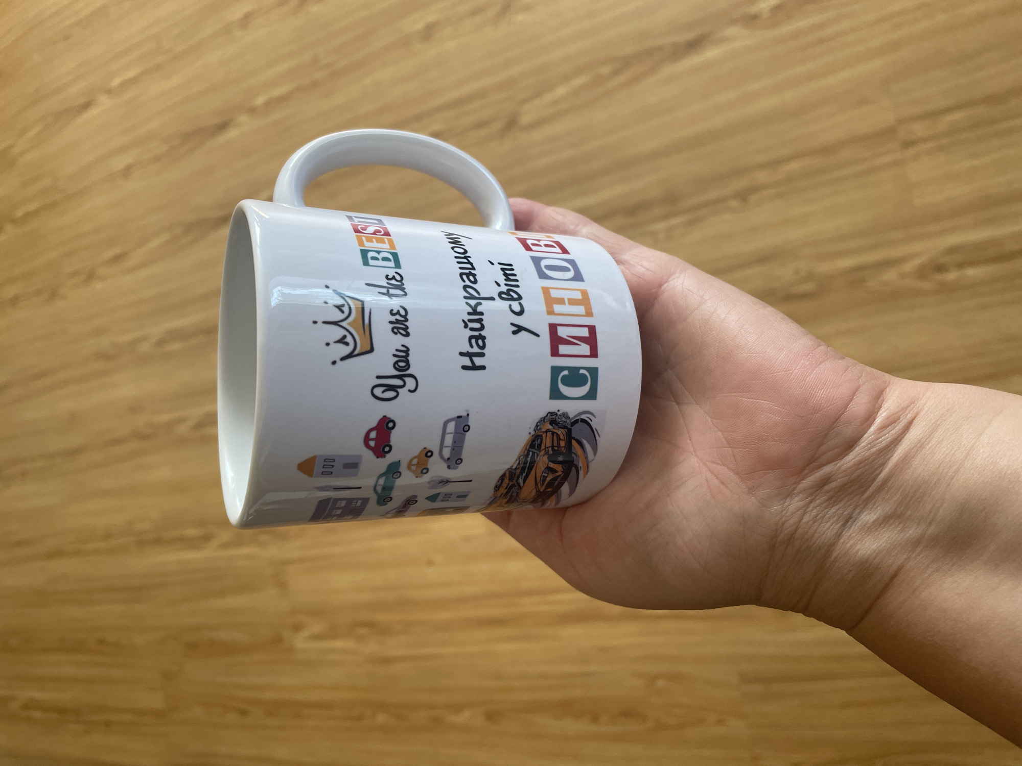 A white ceramic mug is held in a person's left hand against a wooden background. The mug features a cartoonish design of houses and cars, along with the text "You are the BEST" and "Найкращому у світі синові". The text appears to be in English and Ukrainian. The background is a light brown wooden surface with visible grain patterns. The lighting suggests an indoor environment.