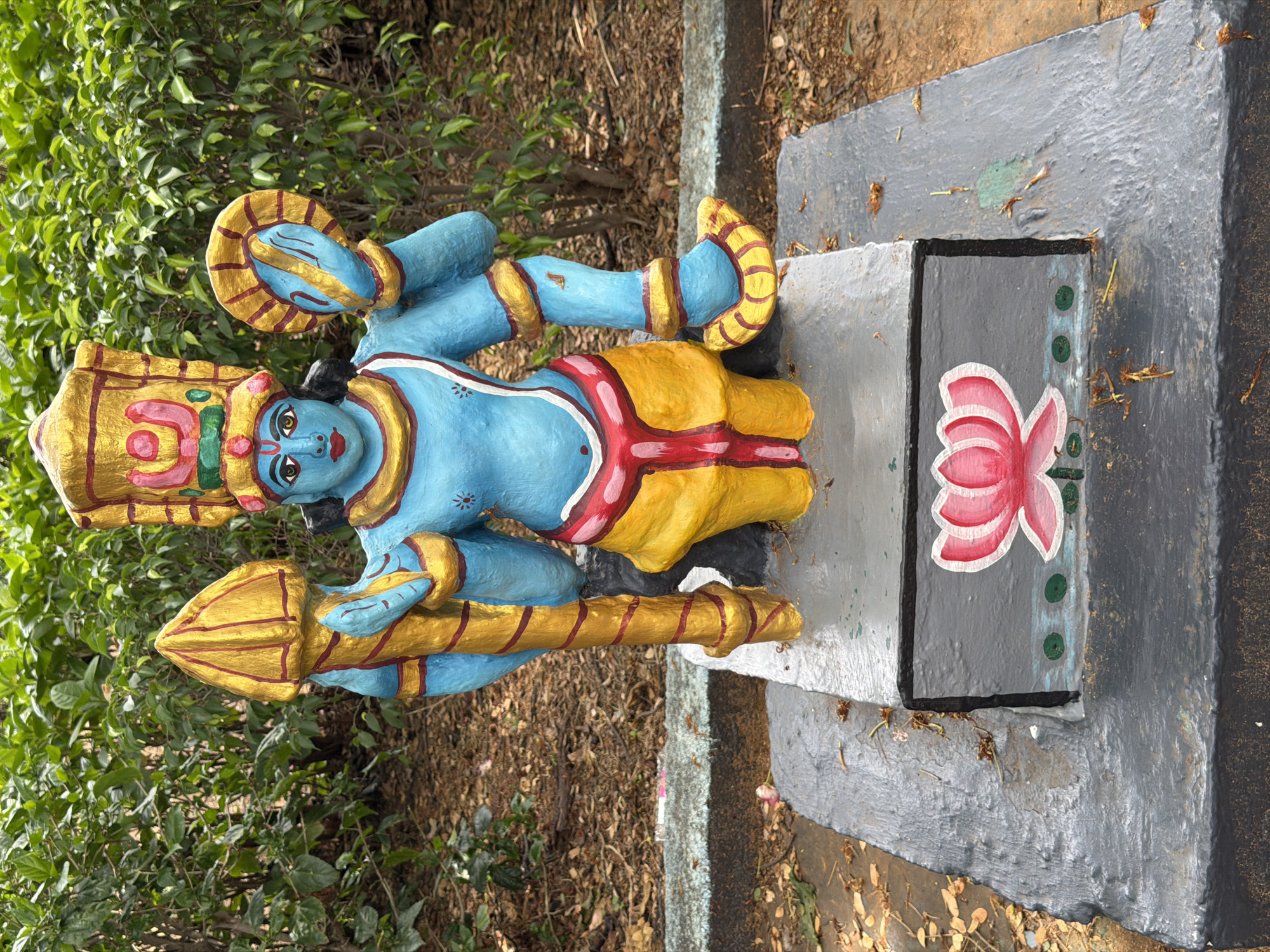 The image shows a brightly painted blue statue of a deity, likely of Hindu origin, standing on a grey, tiered platform. The deity has multiple arms and is adorned with yellow and red clothing, a golden headdress, and golden jewelry. In its hands, it holds a golden mace-like object and a golden disc. The platform itself has a painted pink lotus flower and green dots on its front face. The statue is situated outdoors in a garden setting, with lush green bushes and dry leaves on the ground surrounding it. The overall impression is of a religious or decorative statue in a public space.