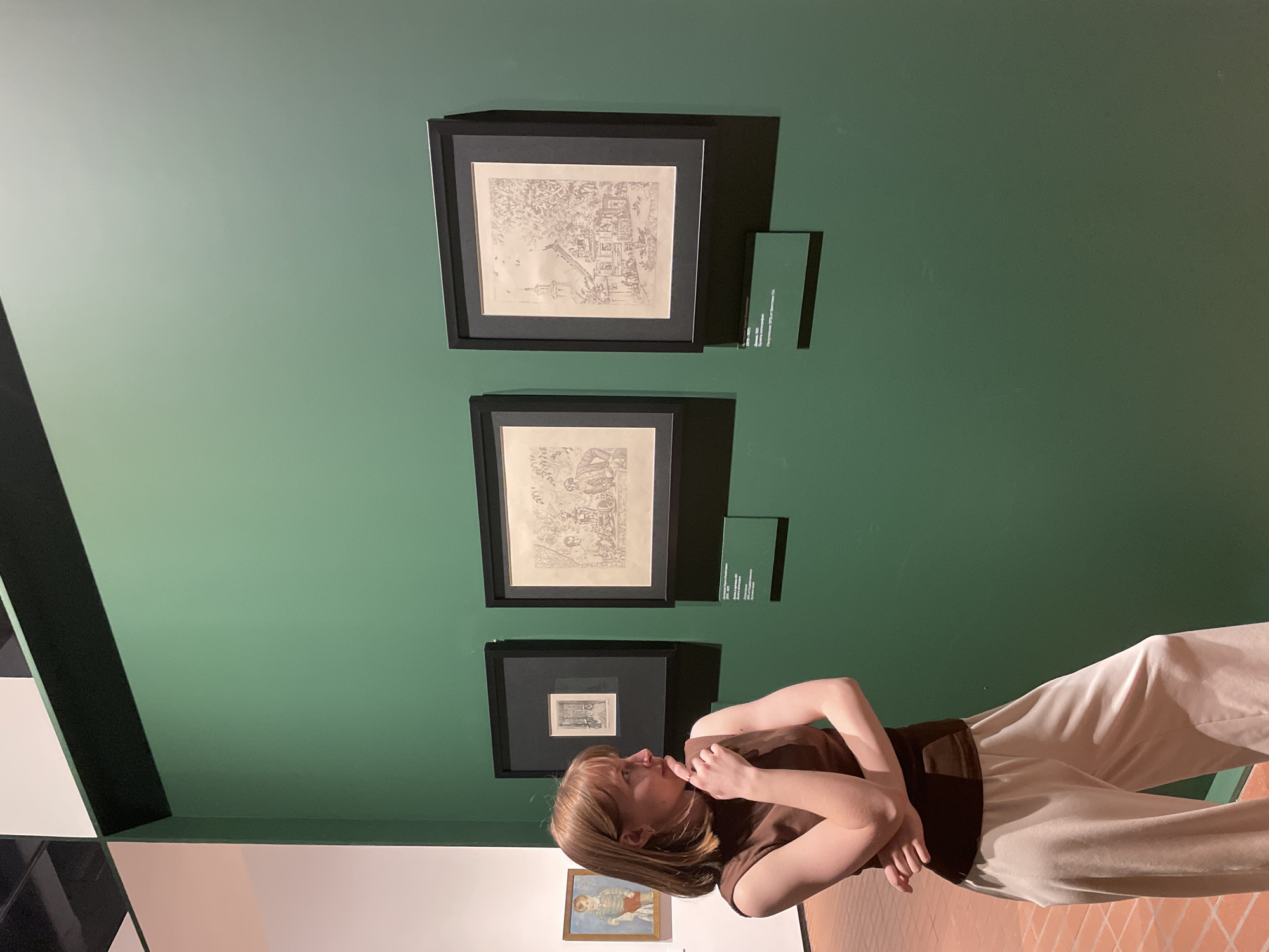 A woman is standing in what appears to be an art gallery in Perm, Russia. She is looking at a set of three framed sketches hanging on a green wall. There is a small informational placard beneath each sketch. A painting of a child hangs further down the wall. The woman is wearing a brown top and light-colored pants. The floor is tiled in a light brown pattern.