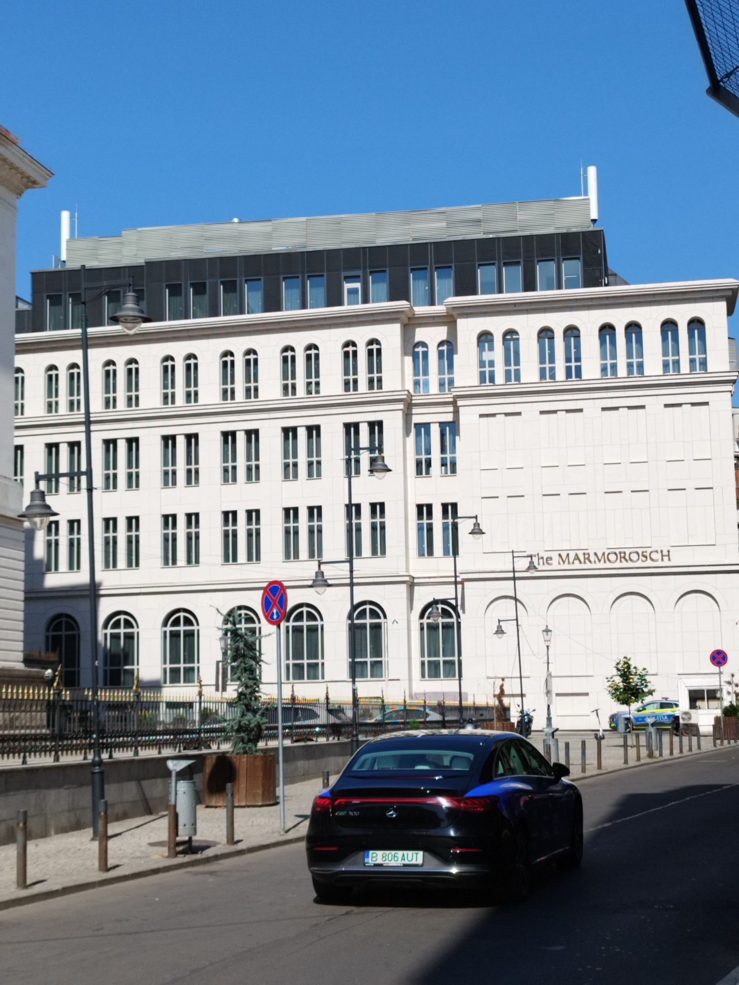 The image captures a street scene in Bucharest, Romania, in front of the Marmorosch Hotel. The Marmorosch is a grand, white stone building with a modern, dark-paneled upper section. The hotel's name is visible in large, dark lettering on its facade.
In the foreground, a sleek black electric car with a license plate that reads "B 806 AUT" is parked or driving away from the camera. The car is casting a shadow on the road. To the left of the car, there's a sidewalk with a black metal fence, a lamppost, and a tall wooden planter with a slender evergreen tree. Further down the sidewalk, a trash can is visible.
On the street, a police car with the word "POLITIA" clearly visible on its side is parked near the curb, accompanied by a person in uniform. Another person is walking in the background. Several lampposts are strategically placed along the street and sidewalk. The sky is a clear, bright blue, indicating a sunny day. The architecture of the Marmorosch Hotel suggests a blend of historical grandeur and contemporary design.