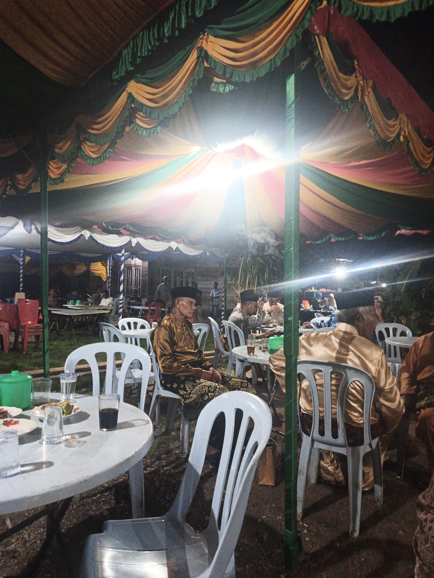 The image depicts an outdoor evening gathering under a large, colorful tent in Pematang Duku, Indonesia. The tent's canopy is adorned with draping fabric in shades of green, yellow, and red, with fringed edges. Several people are seated at white plastic tables, some appearing to be eating or socializing. In the foreground, a man wearing a traditional gold-colored tunic and a black cap is seated. The environment appears to be a courtyard or open space, with visible ground and vegetation in the background. The lighting suggests artificial illumination, possibly from a bright light source above, creating lens flare effects and illuminating the scene. The overall atmosphere suggests a community event or celebration.