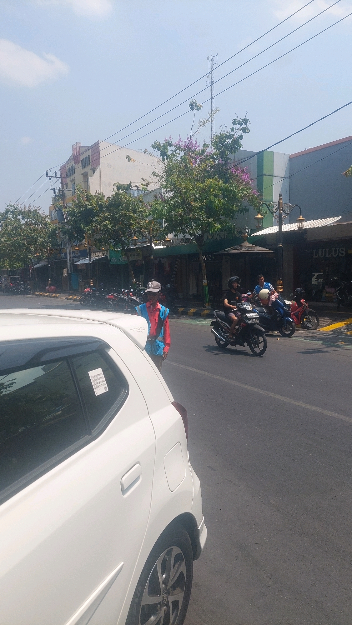 The image depicts a street scene in Madiun, Indonesia, on a sunny day. In the foreground, the rear passenger side of a white car is visible. To the left of the car, a man wearing a red and blue vest over a red shirt and a grey baseball cap stands on the side of the road. He appears to be looking towards the street.
The street itself is paved and appears to be a road with a bike lane marked by green and white paint. Several motorcycles are present on the road. One person is riding a black motorcycle, another person is on a blue motorcycle, and a third person is on a red motorcycle, all moving away from the camera. On the right side of the road, a building with green and white striped accents is visible, with the text "LULUS" partially visible on its facade. Further down the street, other buildings line the road, some with awnings and decorative lighting fixtures. Trees with green foliage and some with purple flowers are planted along the sidewalk. Overhead, power lines stretch across the sky, with a telecommunications tower visible in the distance. The sky is clear with a few wispy clouds.