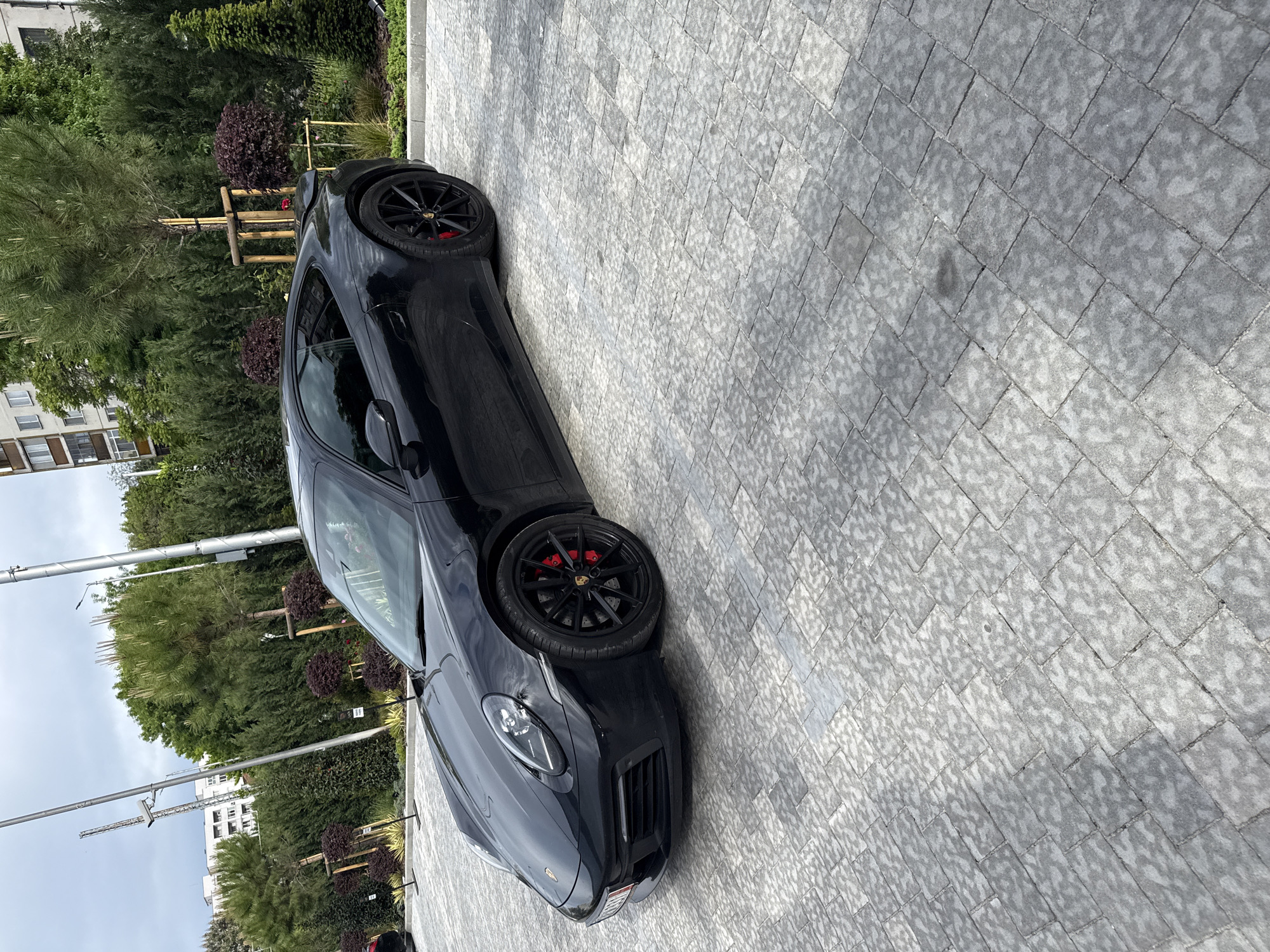 A black Porsche 911 is parked on a gray paved surface. The car is positioned at an angle in the image, showing its front and driver's side. The car has black wheels with red brake calipers. The background features greenery, including trees and bushes. A building is visible in the distance behind the trees. The photo was taken in Plovdiv, Bulgaria.