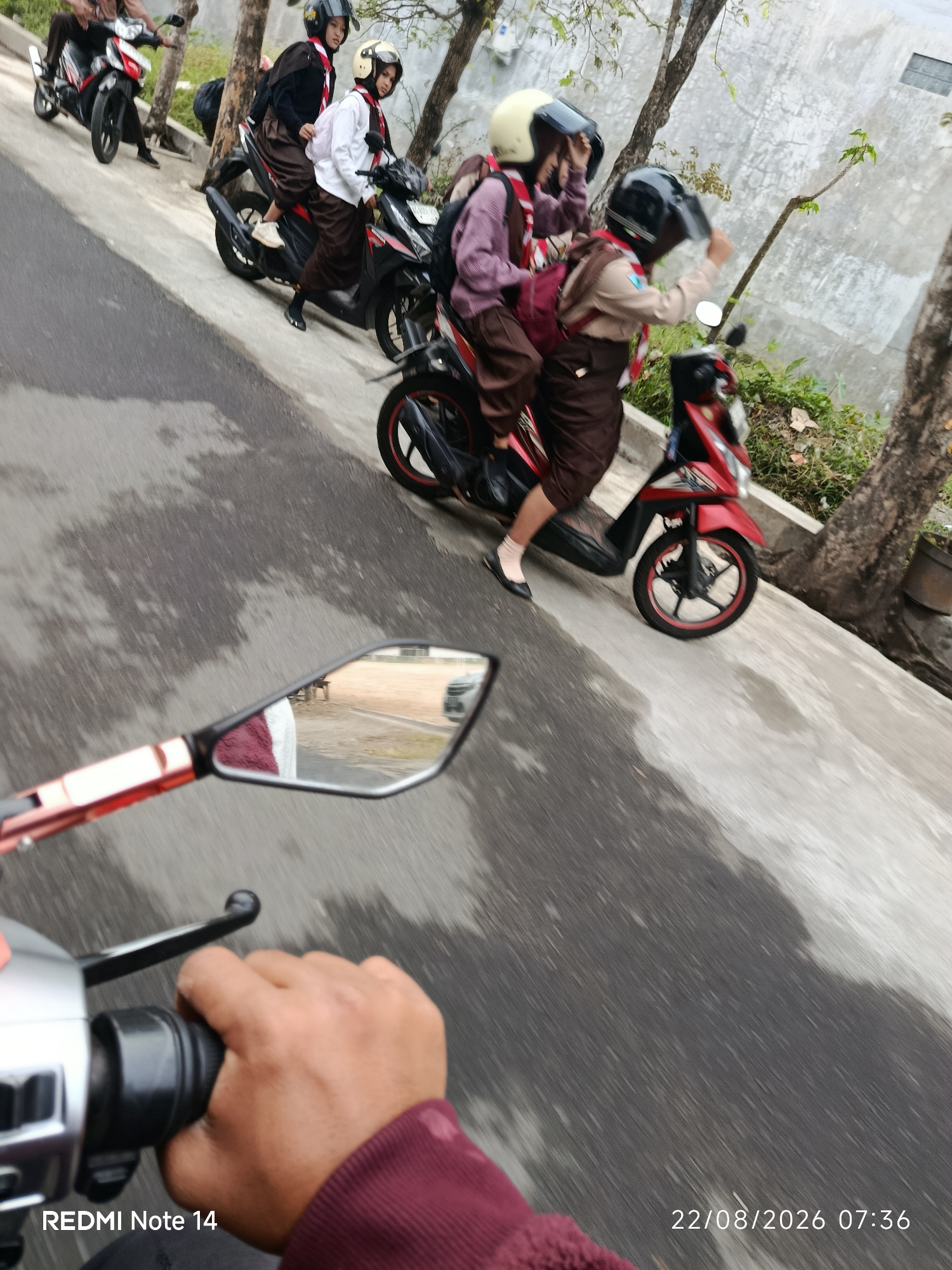 In Mangunsari, Indonesia, on August 22, 2026, at 07:36, three individuals, likely students, are seen riding on two motorcycles. The first motorcycle, a red scooter, carries two individuals in uniform, including a passenger wearing a white long-sleeved shirt and a brown skirt with a yellow helmet, and a driver wearing a brown uniform and a black helmet. The second motorcycle, also a red scooter, has a single individual wearing a brown uniform and a black helmet, possibly driving or as a passenger. A third individual, partially visible, is on a separate motorcycle further in the background. The scene takes place on a paved road next to a concrete embankment lined with trees and sparse vegetation. The foreground features a motorcycle's rearview mirror, reflecting a portion of the road and a bench. The overall image is captured from the perspective of someone on a motorcycle, with a focus on the handlebars and a hand gripping the throttle. The lighting suggests it is daytime, possibly early morning.