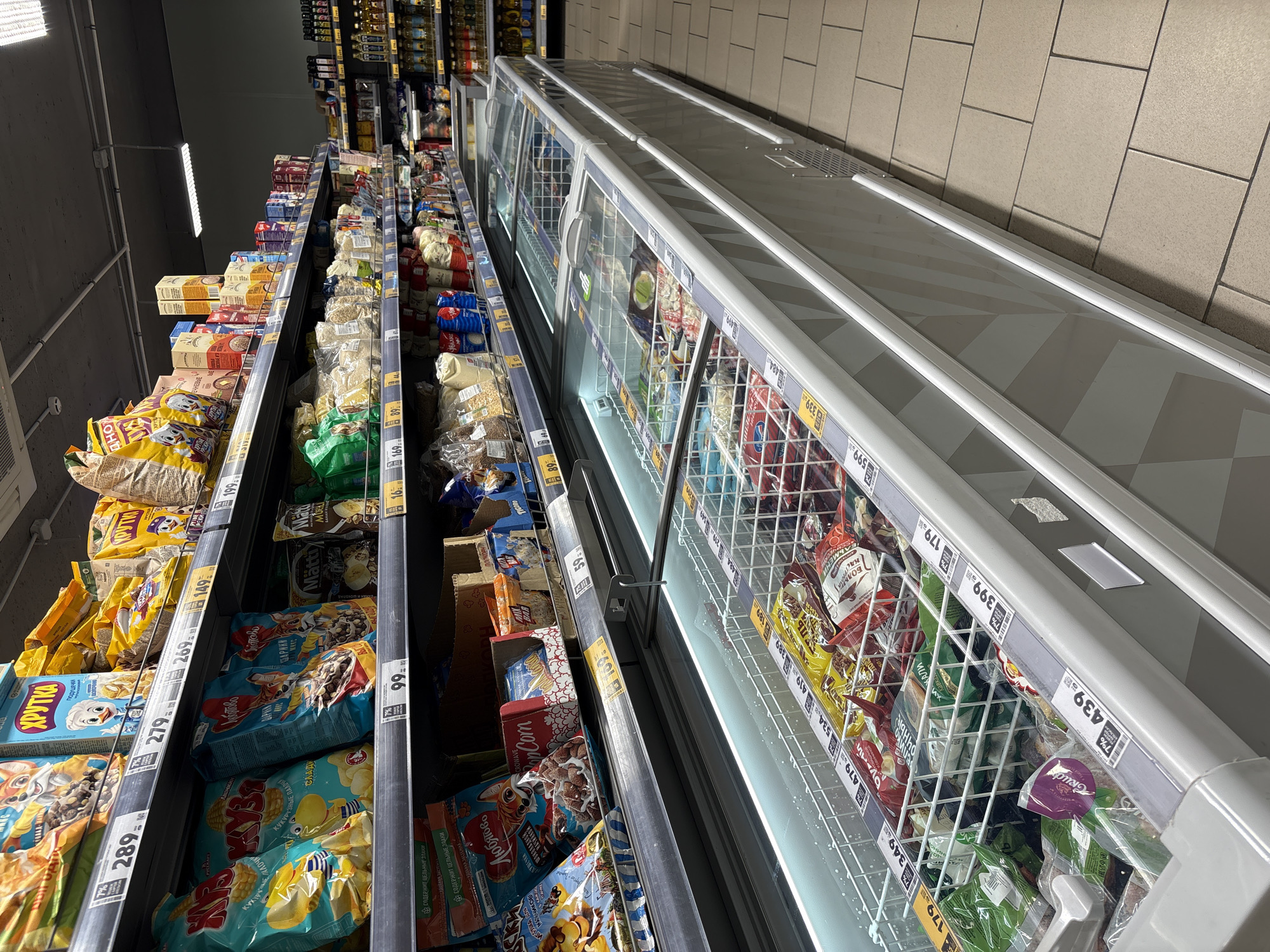 The image shows the inside of a supermarket, likely in Birobidzhan, Russia. The focus is on two aisles. On the left are shelves stocked with boxes and bags of breakfast cereals, including brands like "Khrutka", "Lyubyatovo" and "Matti". Price tags are visible along the shelves. To the right is a freezer section with frozen goods in wire baskets behind glass doors. These goods also have visible price tags. The lighting is typical of a supermarket environment. The aisle floor tiles are visible.