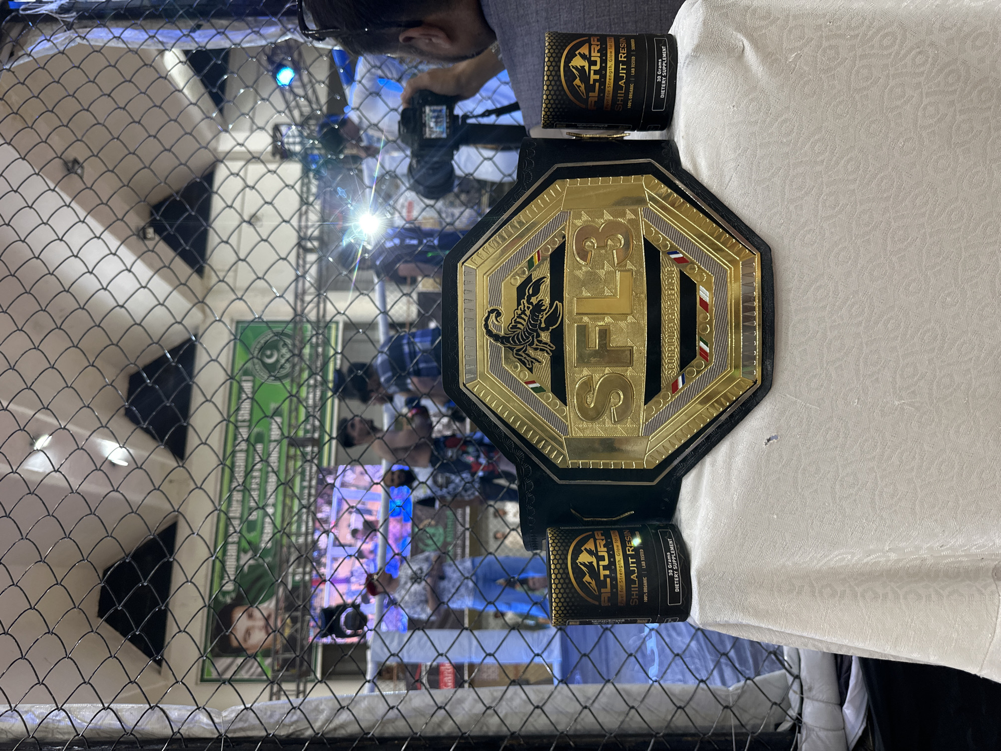 The image captures a mixed martial arts (MMA) championship belt labeled "SFL3" (Scorpion Fighting League) along with two black and gold containers of "ALTURA NATURALS SHILAJIT RESIN," resting on a white tablecloth. The belt is predominantly gold, with a scorpion emblem, lettering, and decorative elements. The background is somewhat blurred, showing what appears to be a live event or crowd behind a chain-link fence, potentially indicating an MMA event. The location is implied to be Quetta City Tehsil, Pakistan. There are also some people in the background taking photographs.