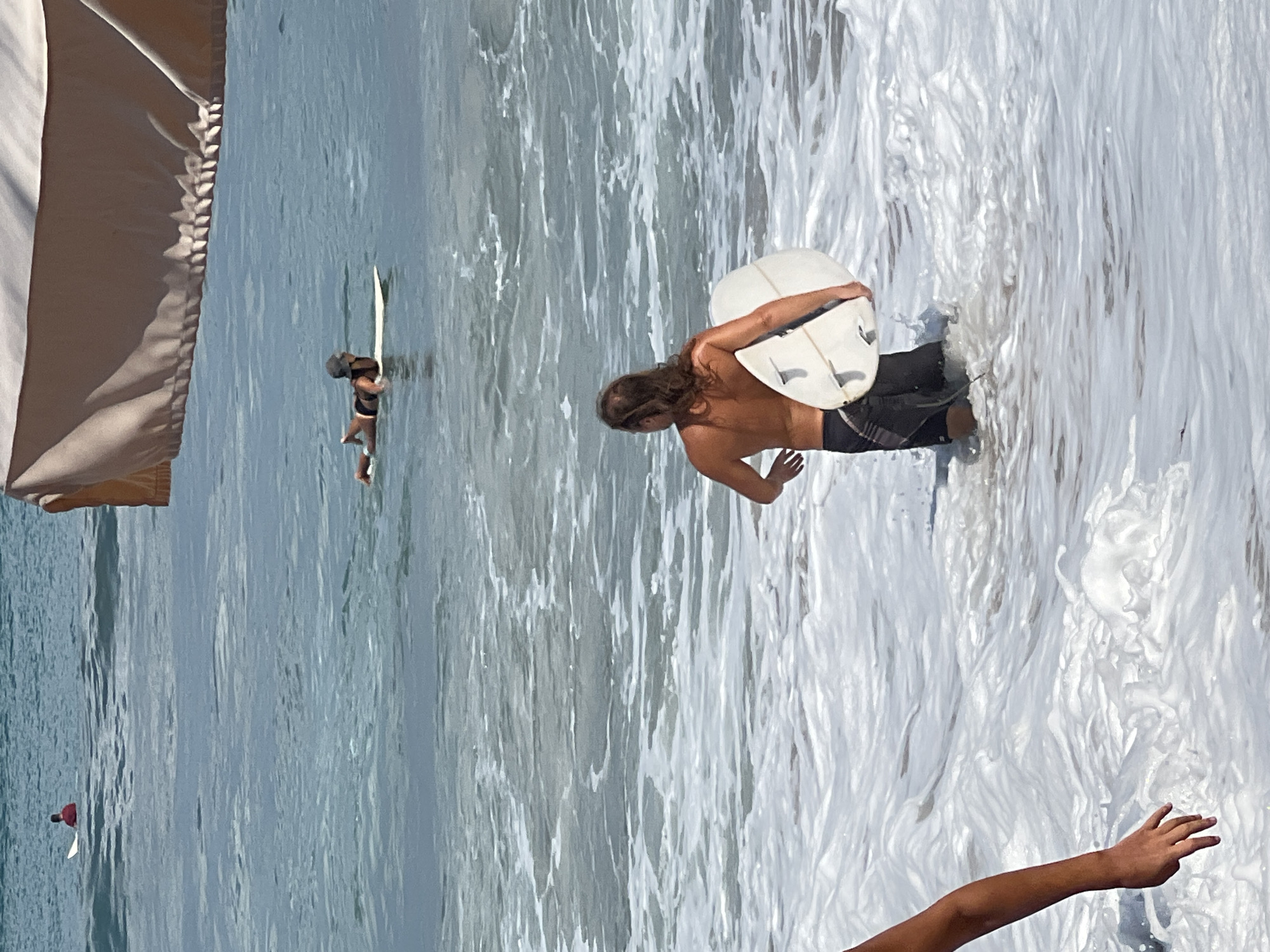 In the ocean waters of Kuta, Indonesia, a person with long, light brown hair, shirtless and wearing dark board shorts, stands waist-deep in foamy waves, holding a white surfboard under their right arm. The surfboard has three fins visible on the underside. To the left, a person's arm and hand are partially visible emerging from the water. Further out in the ocean, another person lies prone on a surfboard, wearing a dark swimsuit and a hat. In the distance, a person in a red shirt sits on a surfboard. The water is a blend of blue and white, with ripples and breaking waves indicating a dynamic sea. A large, light-colored canopy shades the upper right portion of the frame.
