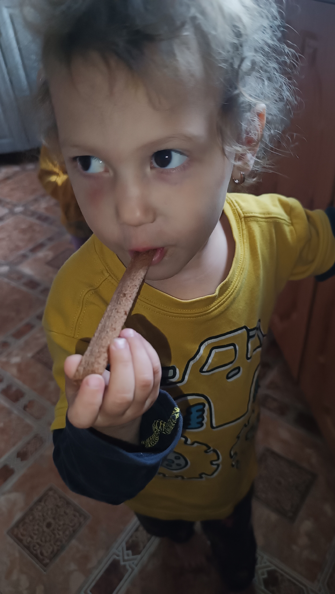 An overhead, medium shot captures a young child with curly blonde hair, wearing a yellow long-sleeved shirt with a dark blue trim and a graphic print, holding a brown, rectangular food item to their mouth and eating it. The child's left eye is looking to the left of the frame, while the right eye is focused forward. A small hoop earring is visible in the child's right ear. The background consists of tiled flooring and what appears to be a wooden cabinet or door. The scene is indoors. The image is not geolocated to Karsha, Russia.