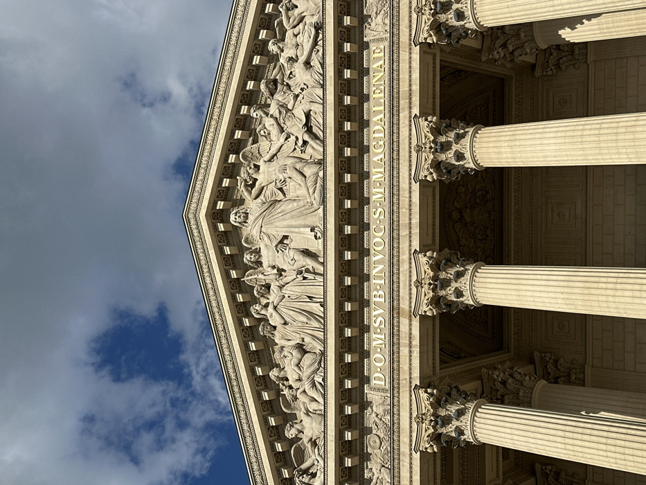 The image displays an upward-angled view of the upper facade of La Madeleine Church, located in Paris, France. The scene is dominated by neoclassical architecture under a partly cloudy sky.
The primary subject is the building's pediment and supporting structure. Six prominent, fluted Corinthian columns, rendered in light-colored stone, are visible at the bottom of the frame, their capitals elaborately carved with acanthus leaves. Above these columns, an entablature features a horizontal inscription in gold capital letters: "D.O.M. SVB INVOC. S. M. MAGDALENAE."
Centered above the inscription is a large triangular pediment containing an ornate bas-relief sculpture. The sculpture depicts numerous human and angelic figures in various poses. A central, robed male figure with outstretched arms dominates the composition, flanked by groups of figures. On the viewer's left, figures, including winged individuals, appear to be rising or looking towards the central figure. On the viewer's right, other figures, some distressed or active, are depicted. The overall action portrayed suggests a dramatic religious event, possibly "The Last Judgment," with figures ascending, interceding, or reacting.
The environment consists of a sky with patches of bright blue visible through scattered gray and white cumulus clouds. Sunlight illuminates the building from the upper right, creating distinct shadows that emphasize the architectural depth and the three-dimensional quality of the pediment sculpture and column fluting.