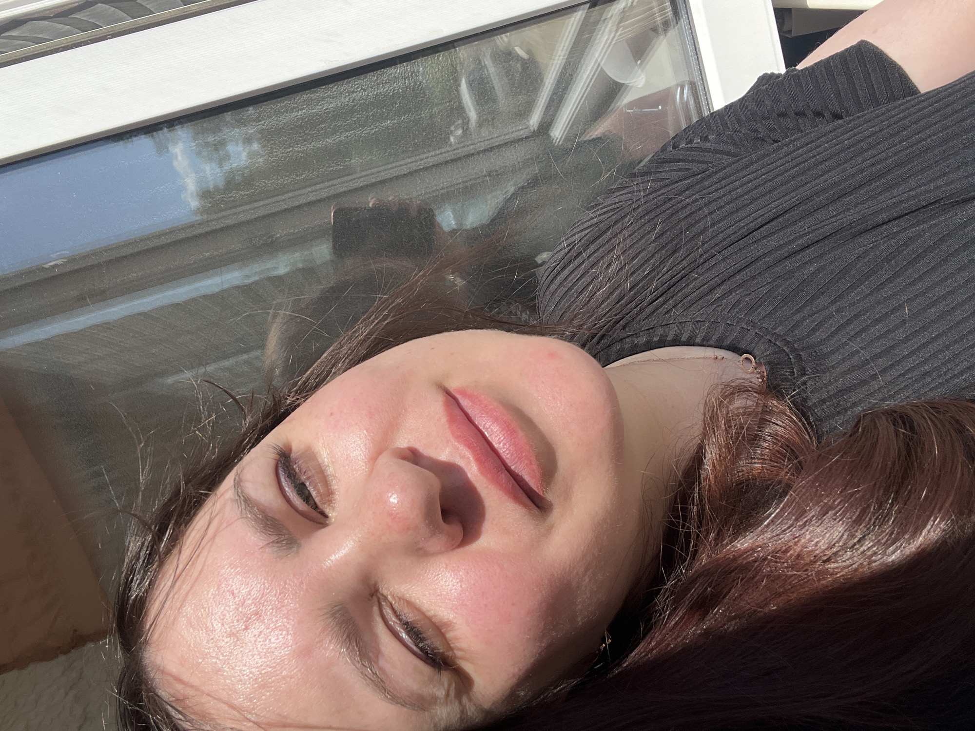 A woman is taking a selfie near a window. She has dark brown hair and is wearing a black shirt. The window is large and shows a reflection of the sky and trees. There is a hint of a building with a white frame visible on the right side of the image. It is a sunny day, as indicated by the bright light and shadows. The image does not contain specific details about the city of Noviy, Russia.