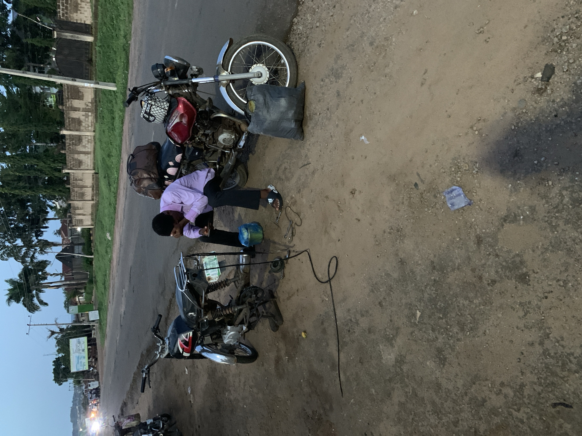 A man is repairing a motorcycle on the side of a road in Gbeleko, Nigeria. The man is wearing a light purple shirt and dark pants. Two motorcycles are visible. One motorcycle is mostly disassembled, and the man is kneeling and working on the exposed engine. The other motorcycle is mostly intact, with baggage strapped to the seat. The road is paved on one side and a dirt shoulder is on the other side. A black bag is laying near the intact motorcycle. A clear plastic wrapper is on the ground on the dirt side of the road. Trees are visible in the background. Buildings are across the road, with distant city lights in the lower left.