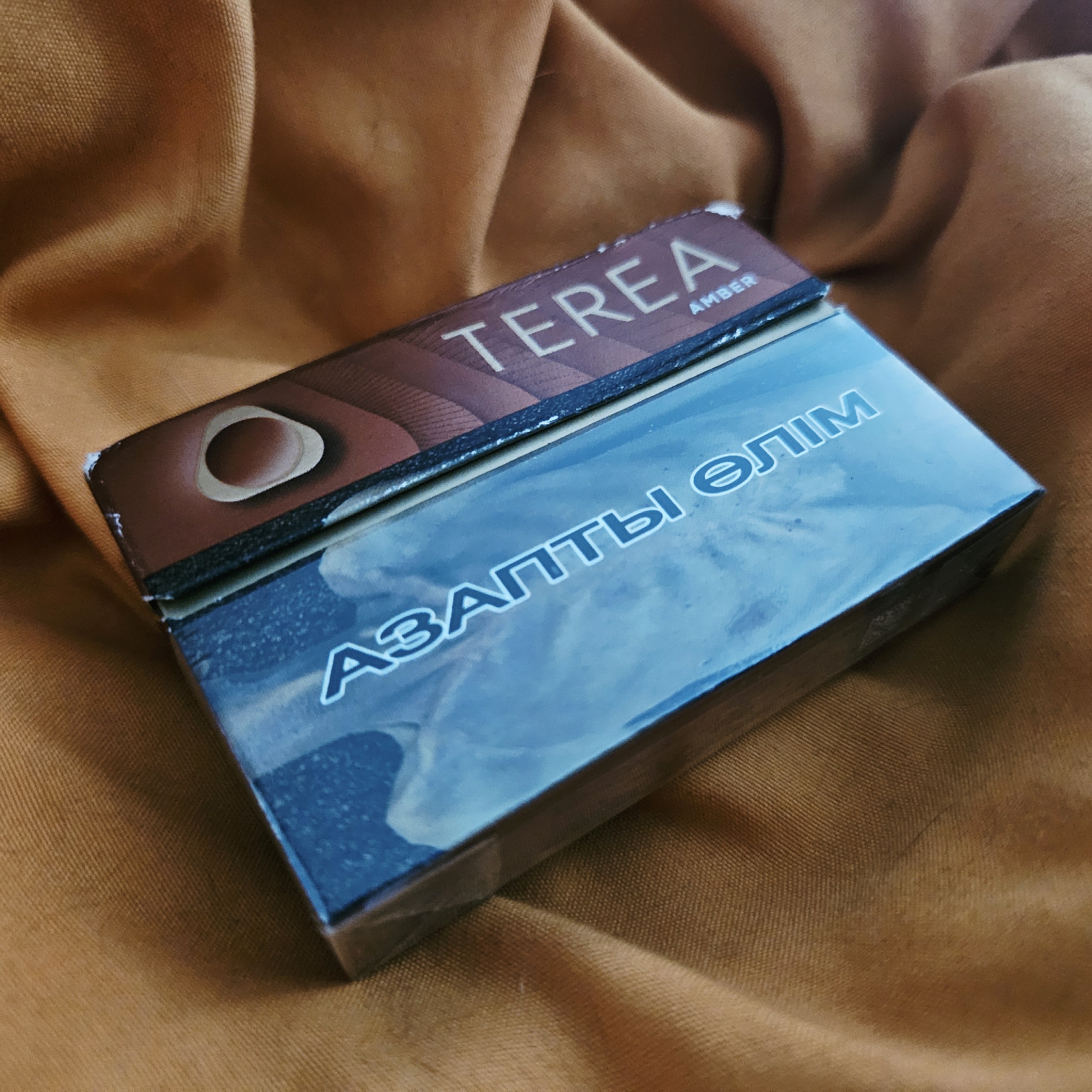 The image shows a close-up of a pack of Terea Amber cigarettes lying on a textured, orange fabric background. The pack is partially wrapped in clear plastic. The top of the pack has a brown design with the text "TEREA AMBER". The main body of the pack features a blue, swirling design with white text in what appears to be Cyrillic script. The lighting suggests an indoor setting. There is no indication of the city of Astana or any specific location within it.