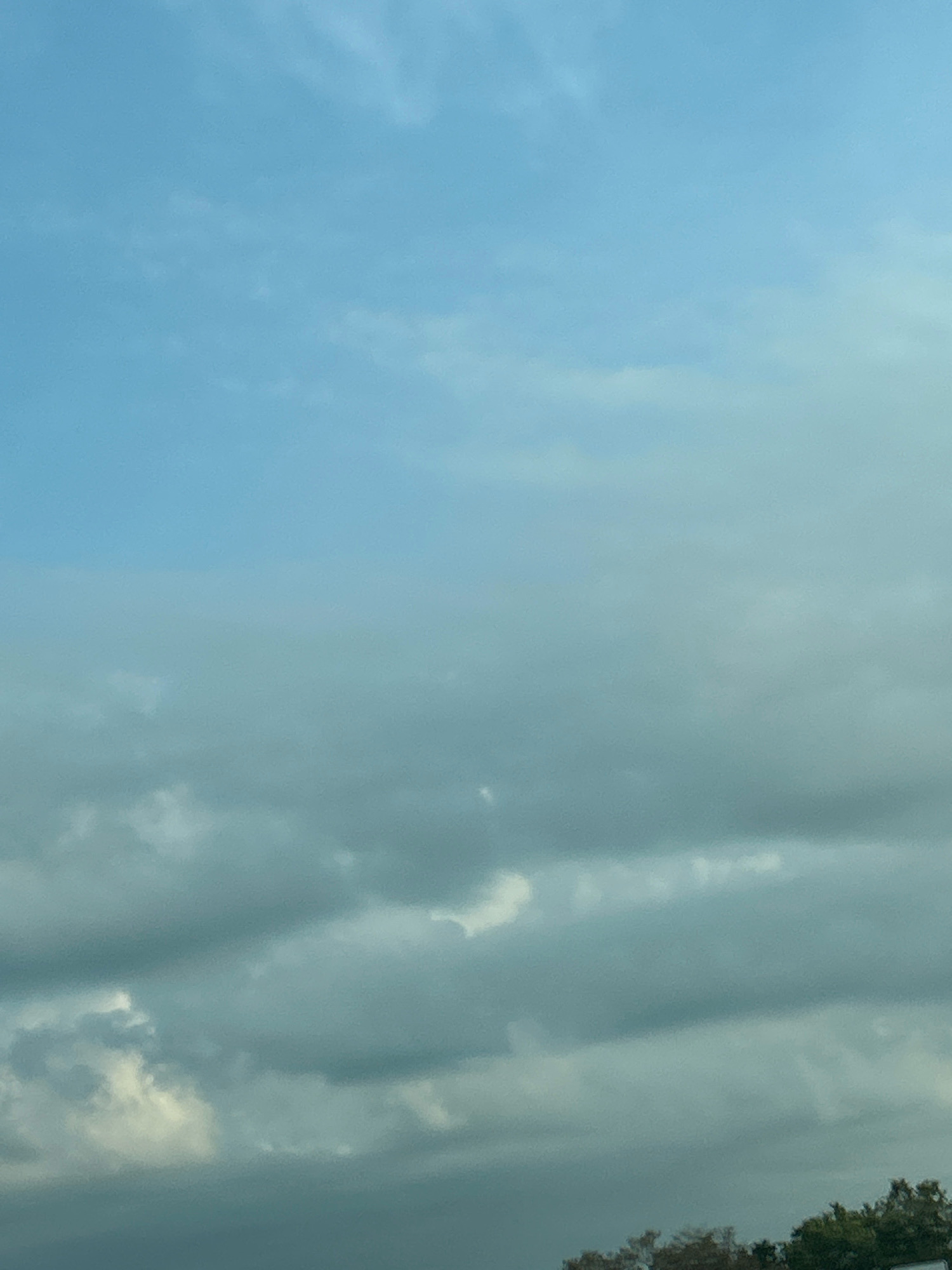 The image depicts a sky with scattered clouds, likely taken from an outdoor location in Robinson Township, United States of America. The sky is a gradient of light blue at the top, transitioning to a lighter, hazy blue and then to a darker, more overcast hue with heavier cloud formations towards the bottom. The clouds are predominantly gray and white, with some areas appearing brighter as if catching sunlight. In the lower portion of the image, a dark line of trees is visible, suggesting the presence of a wooded area or a treeline at the edge of the frame. The overall impression is of a cloudy or partially overcast day.