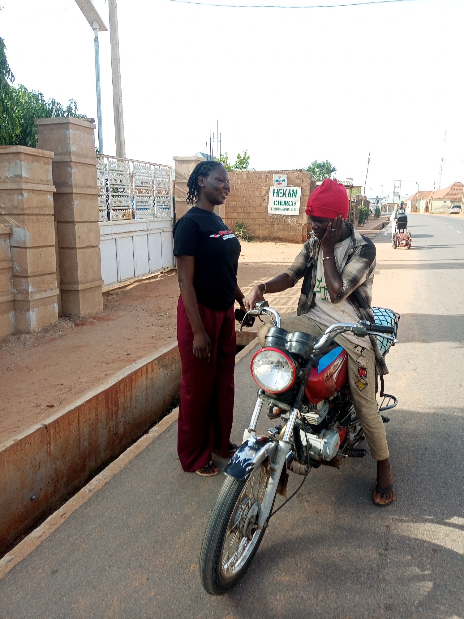 Two individuals are positioned near a motorcycle on a paved street in Gombe, Nigeria. A woman with dark hair is standing to the left of the motorcycle, wearing a black t-shirt and maroon pants. A man wearing a red head covering and a checkered shirt is sitting on the motorcycle. A sign indicating "HEKAN CHURCH, TUMFURE, GOMBE STATE" is visible in the background. In the distance, a person is seated in a motorized wheelchair on the side of the road. Buildings are situated in the background on both sides of the street.