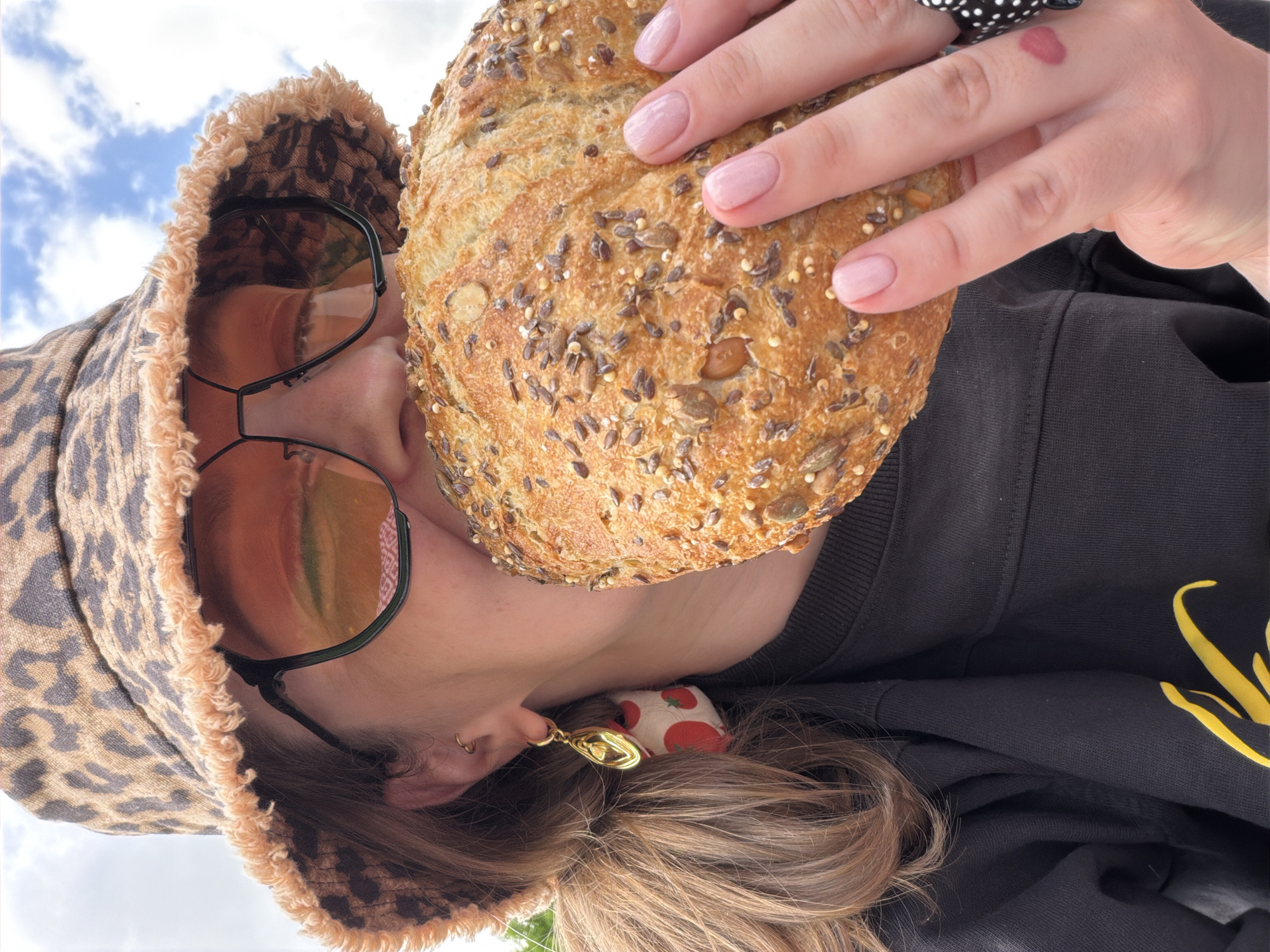 A person, viewed from below, is holding a round loaf of seeded bread close to their face. The person is wearing a black sweatshirt with a yellow design. They also sport a leopard-print bucket hat with fringed edges, and black framed sunglasses with tinted lenses. A strawberry-patterned hair accessory and a gold earring are visible near their ear. The sky, showing blue with white clouds, can be seen in the background. Moscow, Russia is not visible in this image.