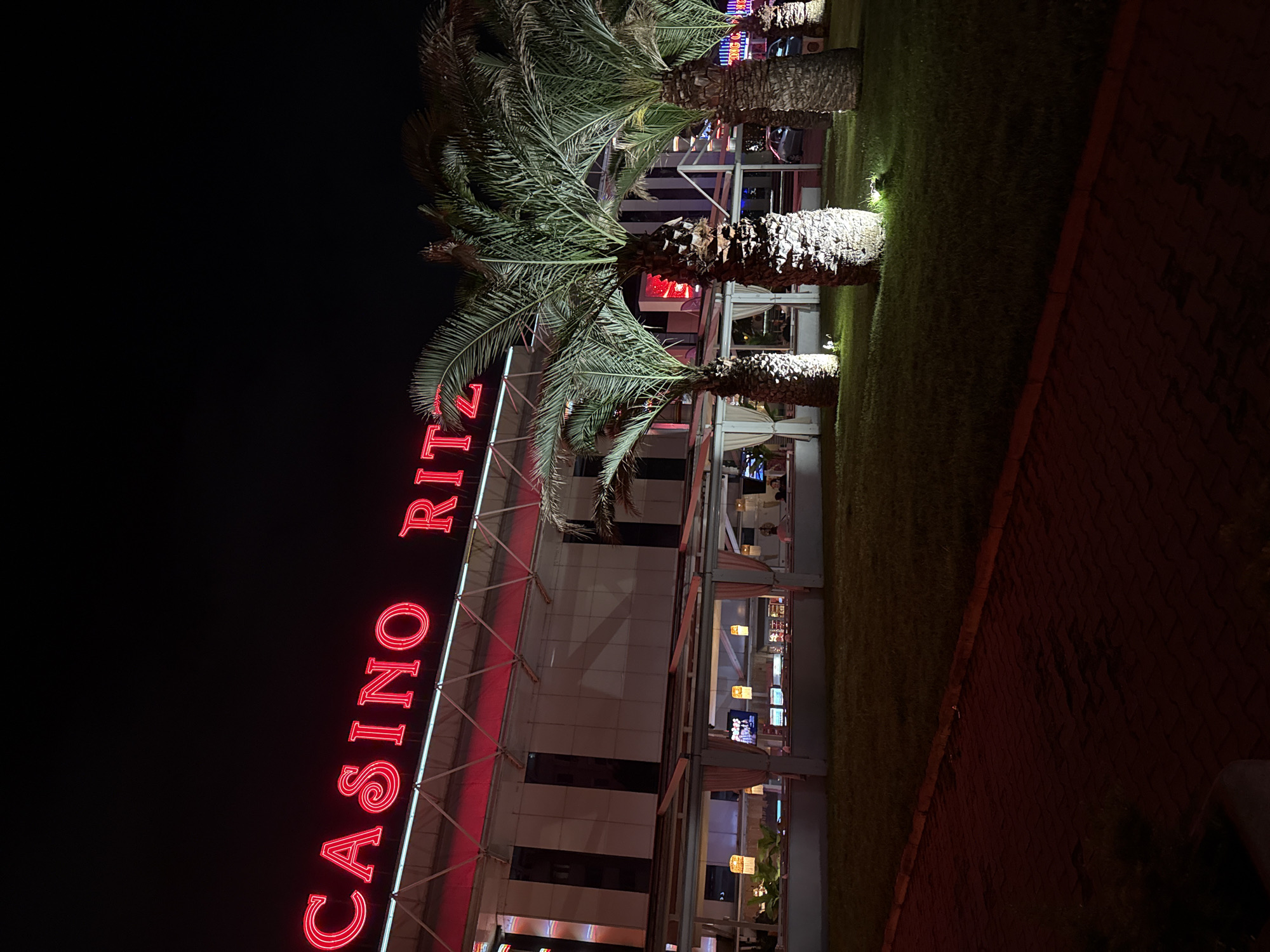 The image captures a nighttime scene featuring the exterior of a building identified by a prominent neon sign. The sign, glowing bright red, spells out "CASINO RITZ." The building has a modern appearance with a glass facade, allowing glimpses of the illuminated interior, where a few indistinct figures can be seen, suggesting people inside.
In front of the building, a row of three palm trees stands tall, their fronds green and visible against the dark sky. The trunks of the palm trees are illuminated from below by spotlights, highlighting their texture. Below the palm trees, there is a neatly maintained strip of green grass, also faintly lit in places, stretching horizontally across the foreground. To the right of the grass, a dark, textured surface, possibly a paved area or sidewalk, is visible, suggesting a pathway or road adjacent to the landscaped area. The overall setting implies an entertainment or resort area.
The time of day is clearly night, as indicated by the dark sky and the reliance on artificial lighting from the neon sign, building interiors, and landscape spotlights. The atmosphere appears vibrant and inviting, characteristic of an evening venue. The palm trees suggest a location with a warm climate.