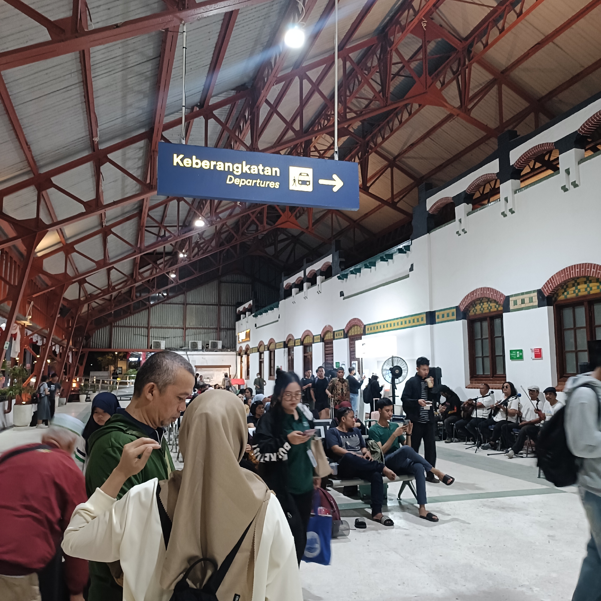 The image captures the interior of an old, grand train station, likely in Semarang, Indonesia, given the signage. A blue sign overhead reads "Keberangkatan Departures" with icons of a person, a bus, and an arrow pointing right, indicating the departure area. The architecture features exposed red steel beams supporting a corrugated metal roof and a white facade with arched windows decorated with brick and colored tiles.
In the foreground, several people are visible. A man in a green jacket, facing away from the camera, is talking to a woman wearing a light brown hijab, whose back is mostly towards the viewer. To the left, a man in a red jacket is partially visible.
In the mid-ground, a group of people are seated on benches, with some standing. A band is playing string instruments, including guitars and violins, to an audience. A young man in a dark shirt and pants is sitting on a bench, looking at his phone. A person in a black jacket and striped shirt is holding a drink and speaking into a microphone. A large standing fan is visible behind the band.
The flooring appears to be light-colored tiles. The overall atmosphere is that of a busy public transportation hub with a performance occurring, possibly to entertain waiting passengers. The lighting is a mix of overhead artificial lights and natural light filtering in from outside.