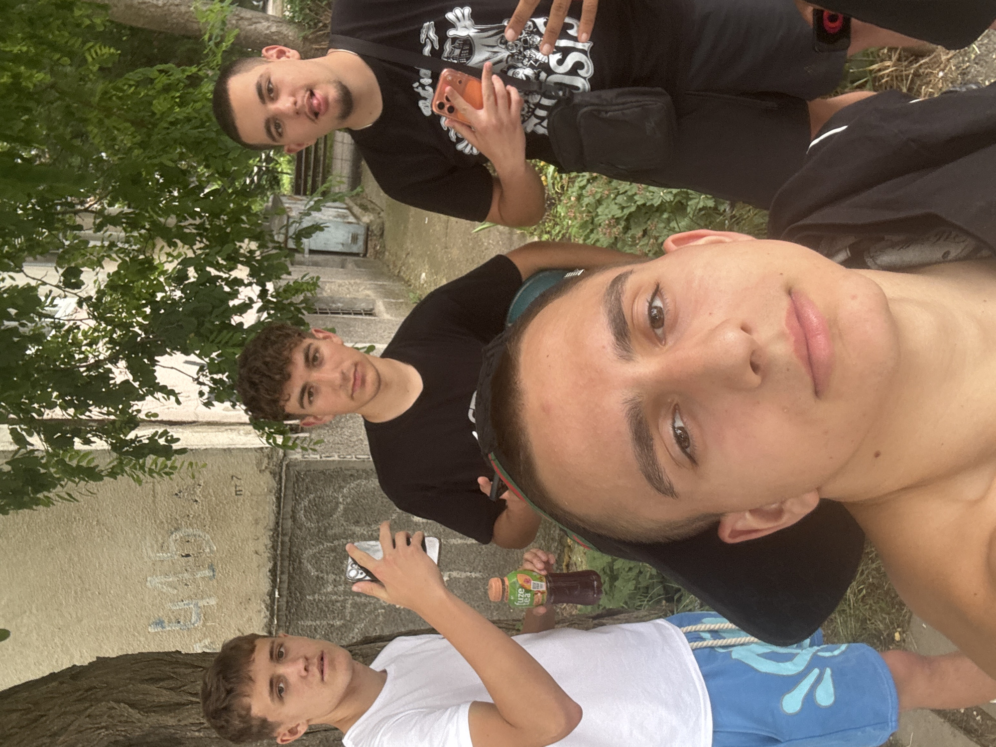 The image is a daytime selfie featuring four young males in an outdoor urban setting, likely in Sofia, Bulgaria.
In the immediate foreground, a young male is seen from the chest up, taking the photo. He has short dark hair and a neutral expression, wearing what appears to be a dark cap and a dark garment draped over his shoulders.
Behind him and slightly to his left, a male with short brown hair wears a white t-shirt and blue shorts with a white drawstring. He holds a silver smartphone in his right hand, pointed towards the left, and a peach-flavored Fuze Tea bottle with an orange label in his left hand. His expression is neutral.
Centered behind the foreground subject, another young male with curly brown hair is visible, wearing a black t-shirt and a dark cap. He looks towards the camera with a neutral expression.
To the right, a young male with short dark hair and a goatee wears a black t-shirt with a white graphic design featuring text and stylized figures, and a black cross-body bag. He holds an orange-cased smartphone in his left hand and is sticking his tongue out towards the camera.
The background consists of an off-white building wall. Faint blue Cyrillic markings "41Б" are visible on the wall, with smaller Cyrillic text "ПТ 7" below. A large tree trunk is on the far left, and abundant green foliage from trees and bushes fills the upper and middle background. The ground visible behind the individuals appears to be unkempt dirt or overgrown earth. The Cyrillic script "41Б" (likely "Block 41B") and "ПТ 7" (potentially an abbreviation for "Ground Floor" or a similar technical designation) are consistent with an address or utility marking in Sofia, Bulgaria.