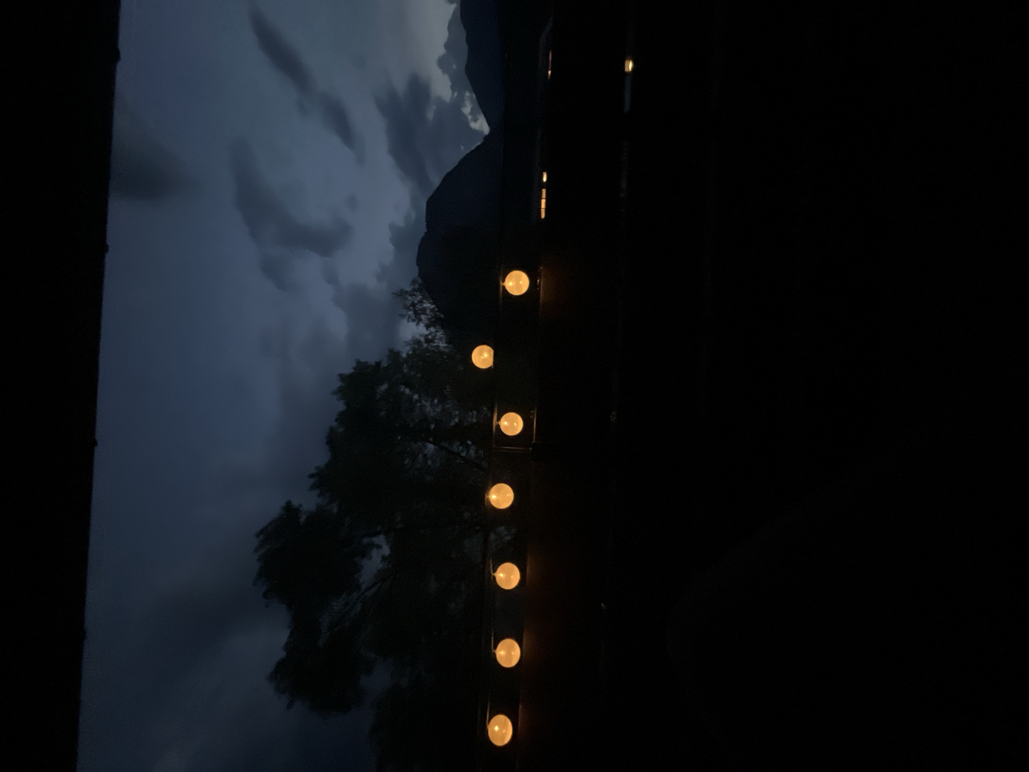 The image captures a twilight scene with a dark, cloudy sky overhead. In the foreground, a line of glowing orange orbs, likely string lights, are visible against the dark silhouette of trees and mountains in the background. The overall mood is somber and slightly ominous, possibly indicating approaching weather or the end of daylight. The location appears to be outdoors, with elements suggesting a rural or mountainous setting rather than an urban one. The specific mention of Garmisch-Partenkirchen, Germany, would require external information not present in the image itself.
