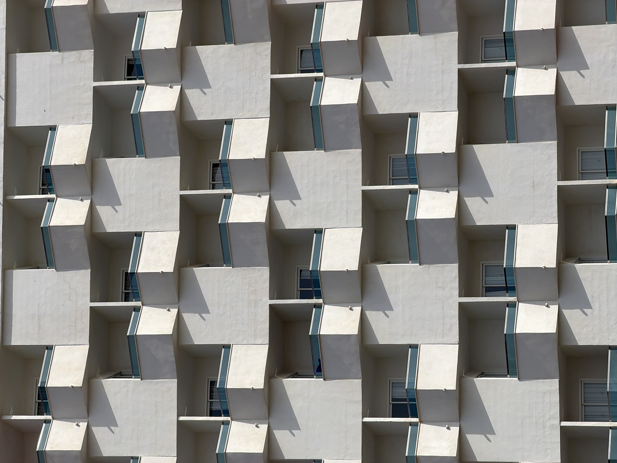 The image shows a repetitive, geometric facade of a building in Magaluf, Spain, likely a hotel or apartment complex. The architecture features a staggered pattern of white balconies, each with a teal-colored railing. The balconies are recessed and project outward, creating a sense of depth and shadow play. Natural light casts distinct shadows on the building's surfaces, highlighting the architectural design. There are no people or objects visible on the balconies, and the scene appears to be an exterior shot of the building's facade. The environment is urban, characterized by the built structure.