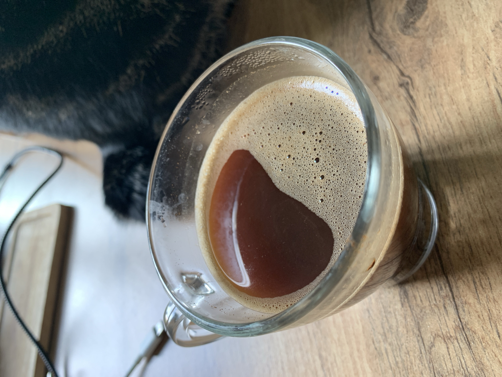 A close-up, high-angle shot shows a clear glass mug filled with dark coffee topped with a thick layer of light brown crema. The crema forms a heart-shaped pattern in the center of the mug, with small bubbles visible throughout. The mug rests on a wooden surface, likely a table, with visible wood grain. In the background, a portion of a dark, furry object, presumably a cat, is partially visible, along with a light-colored cutting board and some cables. The lighting suggests an indoor environment, possibly during the day. The scene is static and there are no people present.
