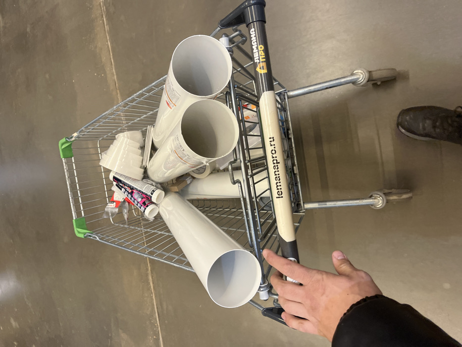 An overhead, first-person perspective image depicts a person pushing a metal shopping cart filled with building materials inside a Lemana Pro (Leroy Merlin Pro) store in Sholokhovo, Russia. The person's right hand, wearing a dark sleeve, grips the cart's light-colored handle, which displays "lemanapro.ru" and "ЛЕМАНА ПРО" (Leroy Merlin Pro) branding. The person's left foot, shod in a dark shoe, is visible on the light gray, industrial-style floor. The cart contains several large, light gray PVC pipes, multiple tubes of sealant or adhesive with white, red, and black labels, and a stack of white plastic cups or containers. Other indistinct items are also present within the cart.