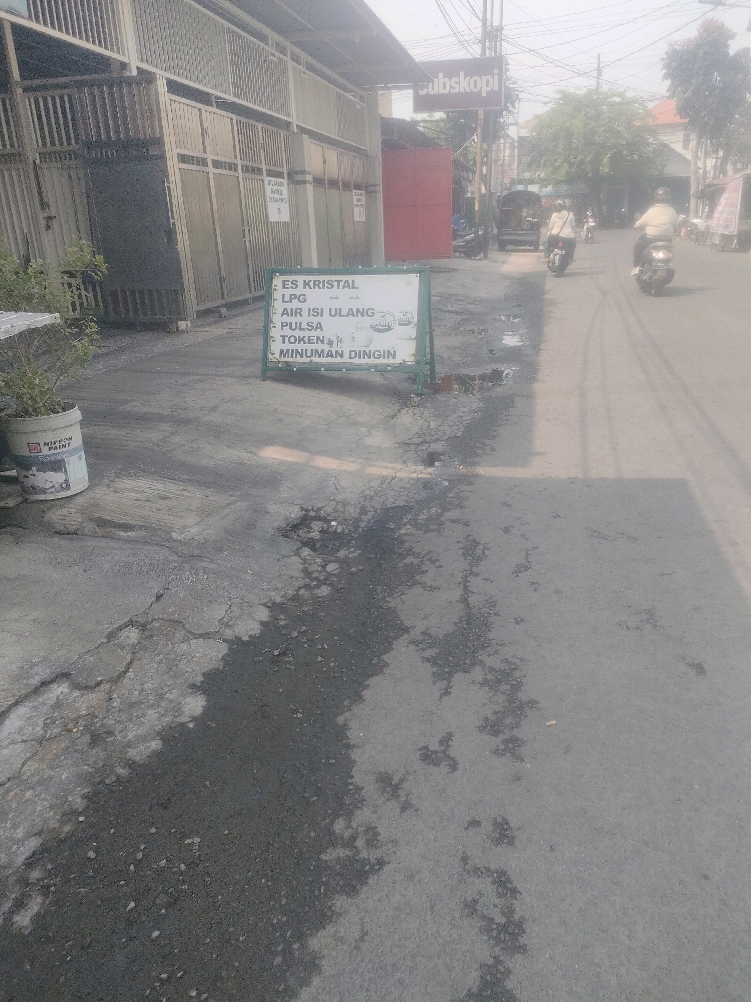The image depicts a street scene in Surabaya, Indonesia, characterized by its hazy atmosphere, suggesting either pollution or a dusty environment. In the foreground, a sign displays a list of items for sale: "ES KRISTAL," "LPG," "AIR ISI ULANG," "PULSA," "TOKEN," and "MINUMAN DINGIN." Adjacent to the sign is a white paint bucket from "NIPPON PAINT." The street itself is asphalt, showing signs of wear and tear with patches of moisture or oil stains. Further down the street, two individuals are riding motorcycles, and a three-wheeled vehicle is also visible. Buildings line the street, with a prominent sign reading "ubskopi" on one of them. The overall impression is that of a typical urban street in Indonesia, possibly near a local shop or minimarket.