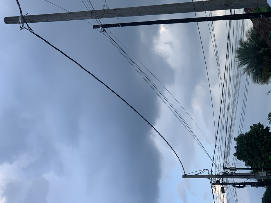 The image captures an outdoor scene dominated by a dense network of utility wires and poles against a dramatic, cloudy sky. No people are visible.
The scene features several concrete utility poles, with one prominent pole on the far right supporting a multitude of tangled black wires and possibly some utility boxes or equipment. Another pole is partially visible on the far left. A thick, dark cable runs diagonally across the frame from the top left to the bottom right, appearing to sag slightly. Numerous other thinner wires crisscross the sky, creating a complex web.
The sky itself is a notable feature, presenting a distinct contrast. The left side of the frame is covered by a large, dark, uniform mass of clouds, suggesting heavy overcast conditions or an approaching storm. In stark contrast, the right side of the sky shows lighter, more broken clouds with patches of brighter sky, possibly indicating sunlight attempting to penetrate or a clearing weather pattern. This creates a clear boundary between the dark and light cloud formations.
Vegetation is visible on the right side of the image, including the fronds of a palm tree near the top right corner and the green canopy of other leafy trees towards the bottom right. The overall impression is that of a bustling urban or suburban environment, characteristic of regions like Metro, Indonesia, where such dense overhead utility infrastructure is common. The time of day appears to be daytime, possibly late afternoon, given the varying light in the sky. There is no visible text in the image.