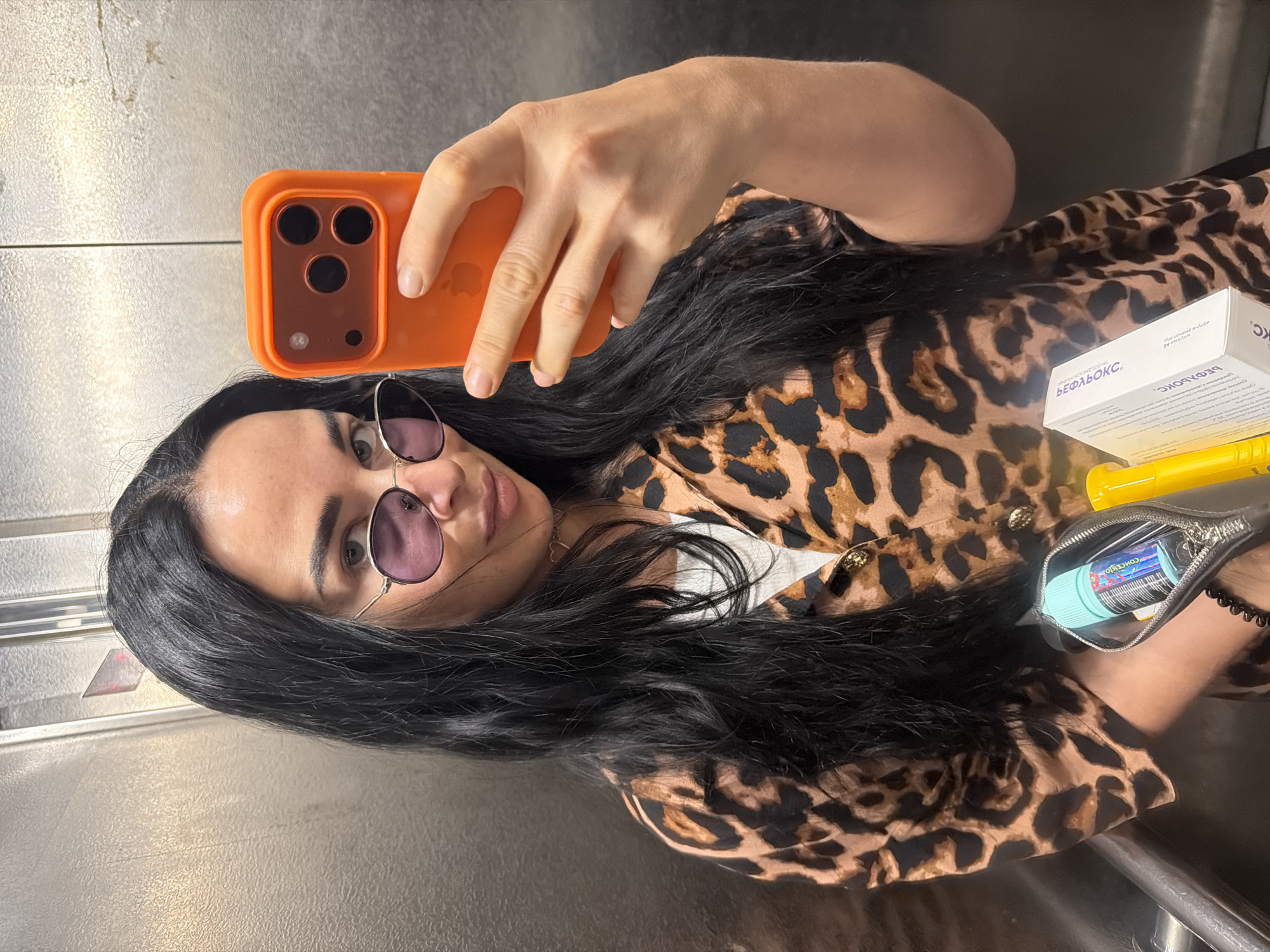 A woman with long, dark hair and purple-tinted sunglasses is taking a selfie in an elevator with metallic walls. She is wearing a leopard-print button-up shirt over a white top. In her right hand, she holds an orange smartphone, and in her left hand, she is holding a small black pouch that contains a blue bottle of vape liquid and a yellow object. A white box with Cyrillic text is also visible.