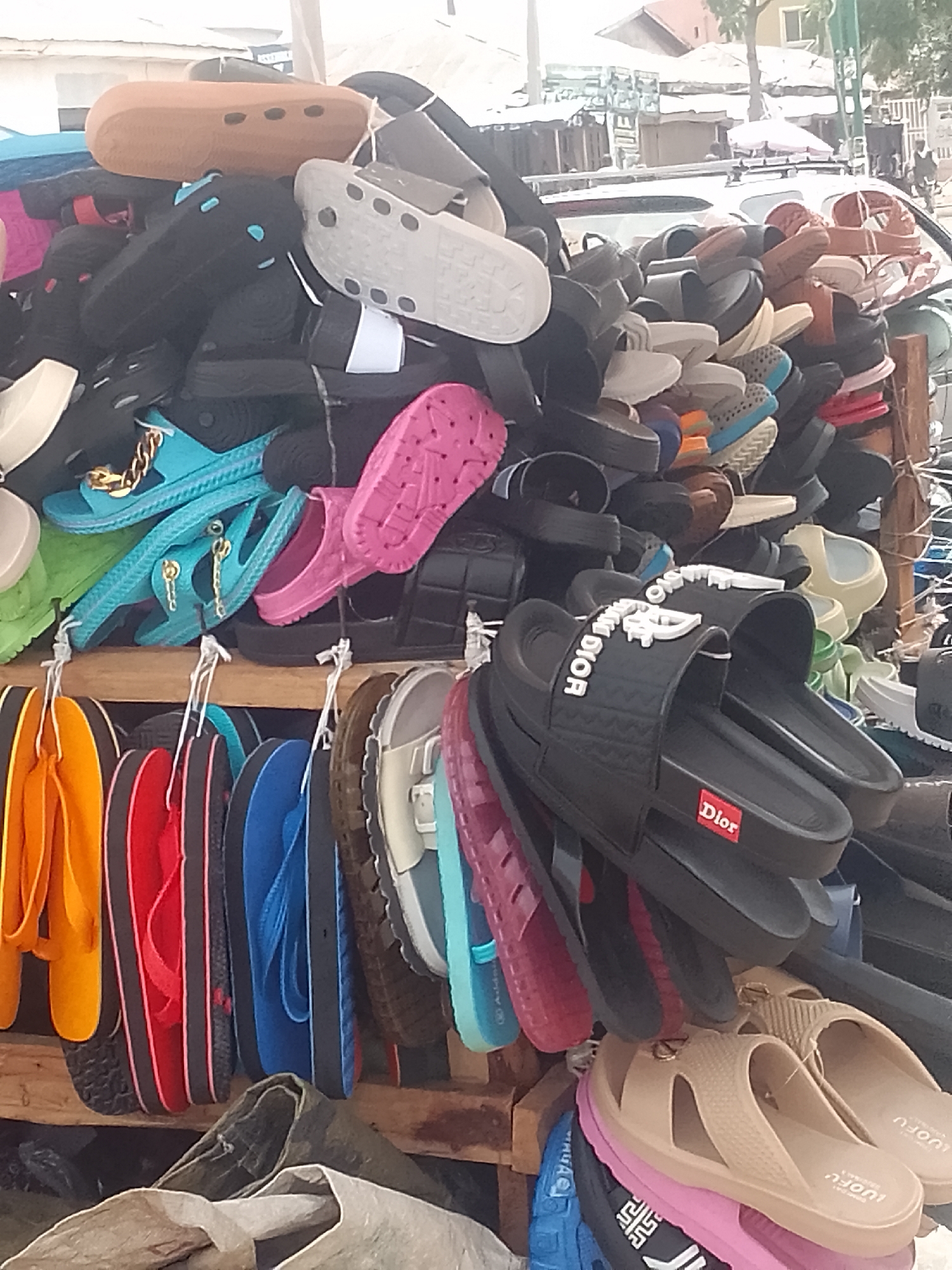 A street vendor in Pantame, Nigeria displays a large assortment of footwear. Sandals, flip-flops, and slide sandals, in a variety of colors, are stacked high and hung from racks. A few pairs of black "Dior" slides are visible. In the background is a road with a few cars. Buildings are seen in the far background.