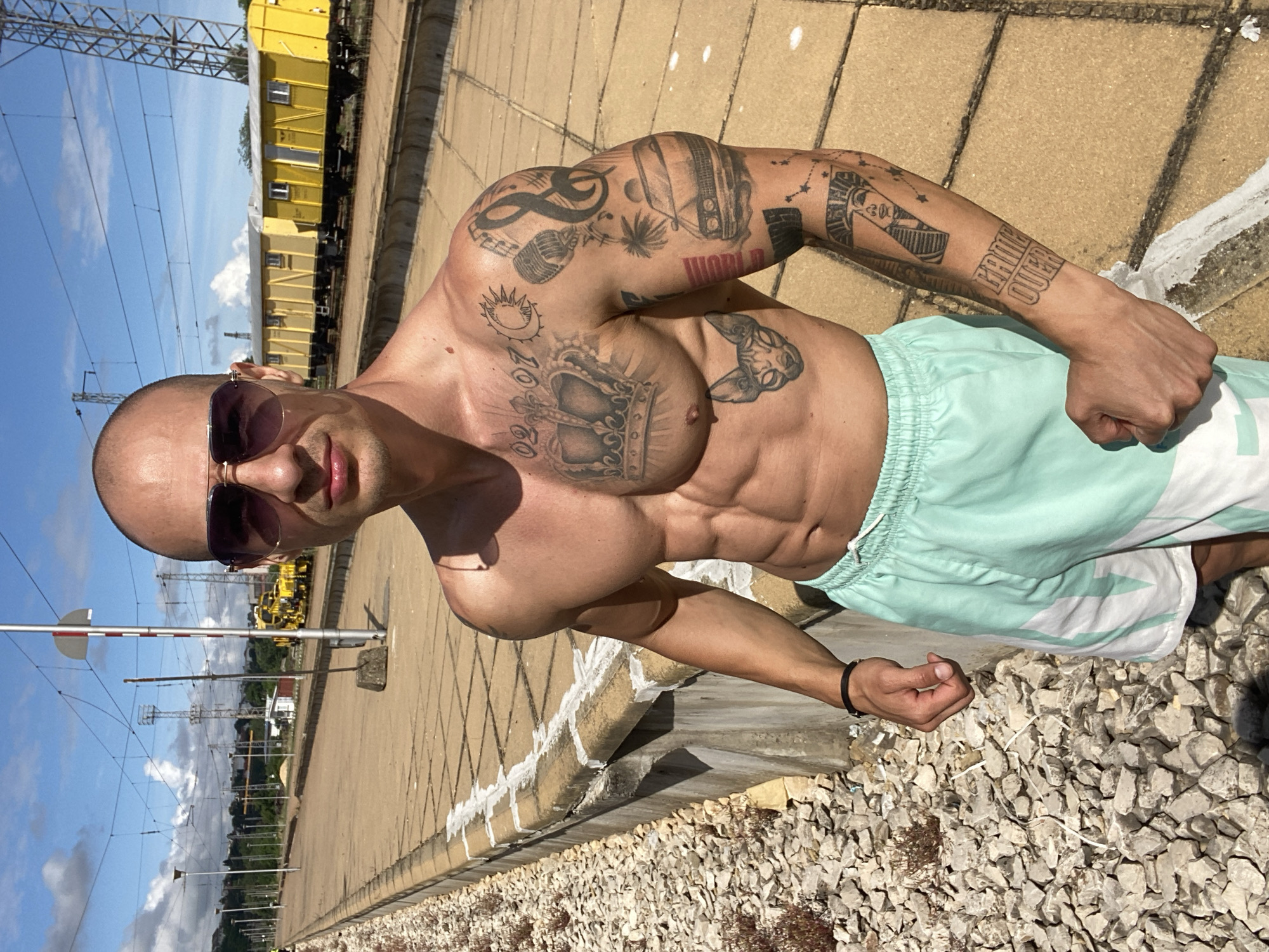 A bald, muscular man wearing aviator sunglasses and light green and white patterned shorts stands on a train platform. He has tattoos on his chest and arms. The platform is made of concrete tiles and is lined with rocks and a low concrete wall. In the background, yellow train cars are visible, along with overhead power lines and distant buildings. The sky is blue with scattered clouds.