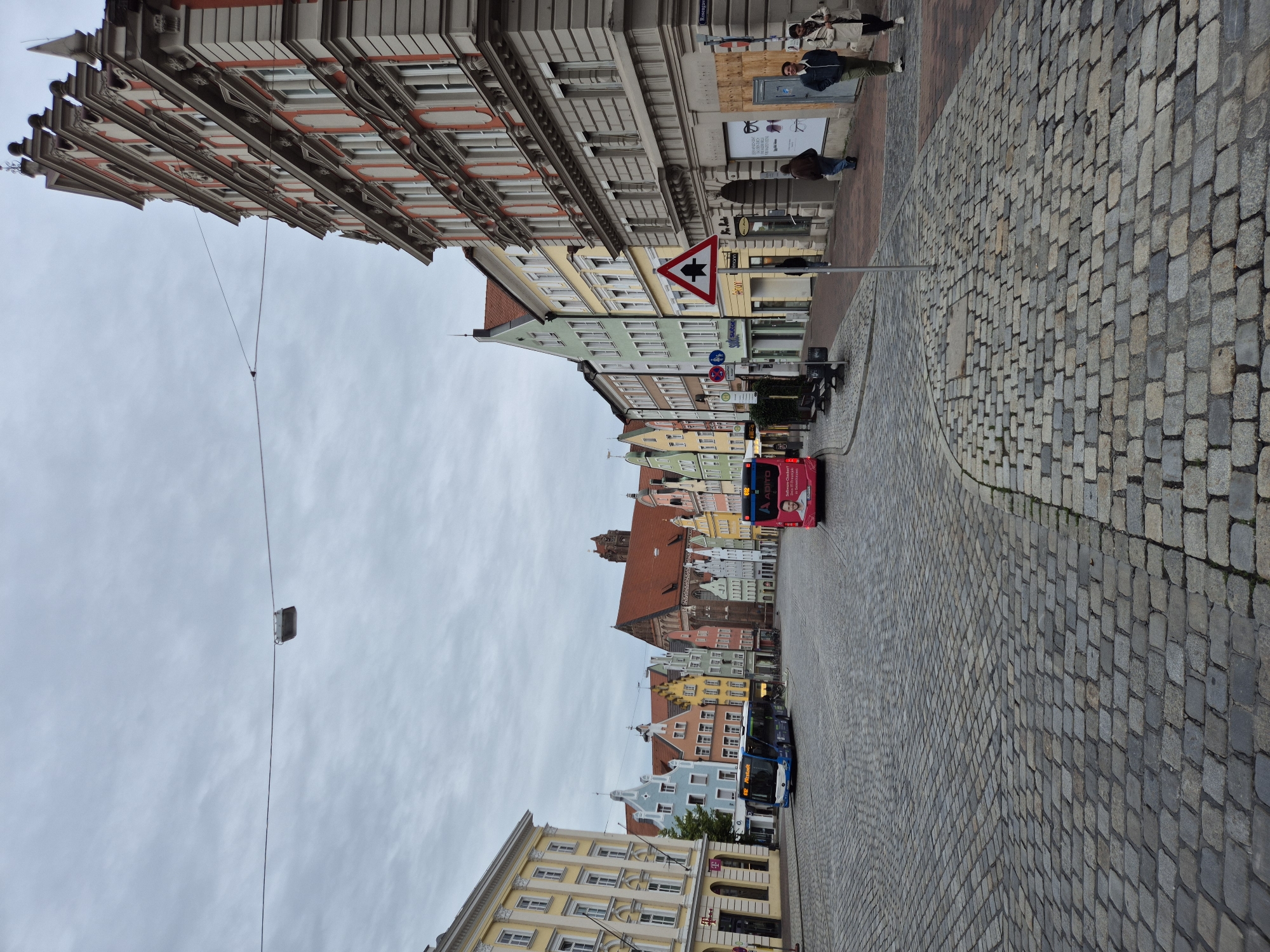 The image shows a view of the Altstadt, the historic old town of Landshut, Germany, under an overcast sky. The cobblestone street, the Altstadt, is lined with colorful, multi-story buildings in a mix of architectural styles, many with pitched roofs. On the left, a modern yellow building stands out. In the center of the street, two buses are visible: a dark blue bus in motion and a bright pink bus parked.
Several people are present. Two individuals are walking on the right sidewalk, a man in a dark jacket and green pants and a woman in a cream-colored coat. Another person, wearing a brown jacket, is walking further down the sidewalk. A figure in a dark jacket is seen walking on the sidewalk in the middle ground.
Various street furniture and signs are present, including lampposts, benches, traffic signs, and shop signage. A triangular warning sign with a pedestrian symbol is prominently displayed. Some shop windows are visible, one displaying eyewear. The overall impression is of a quiet, historic European street scene.