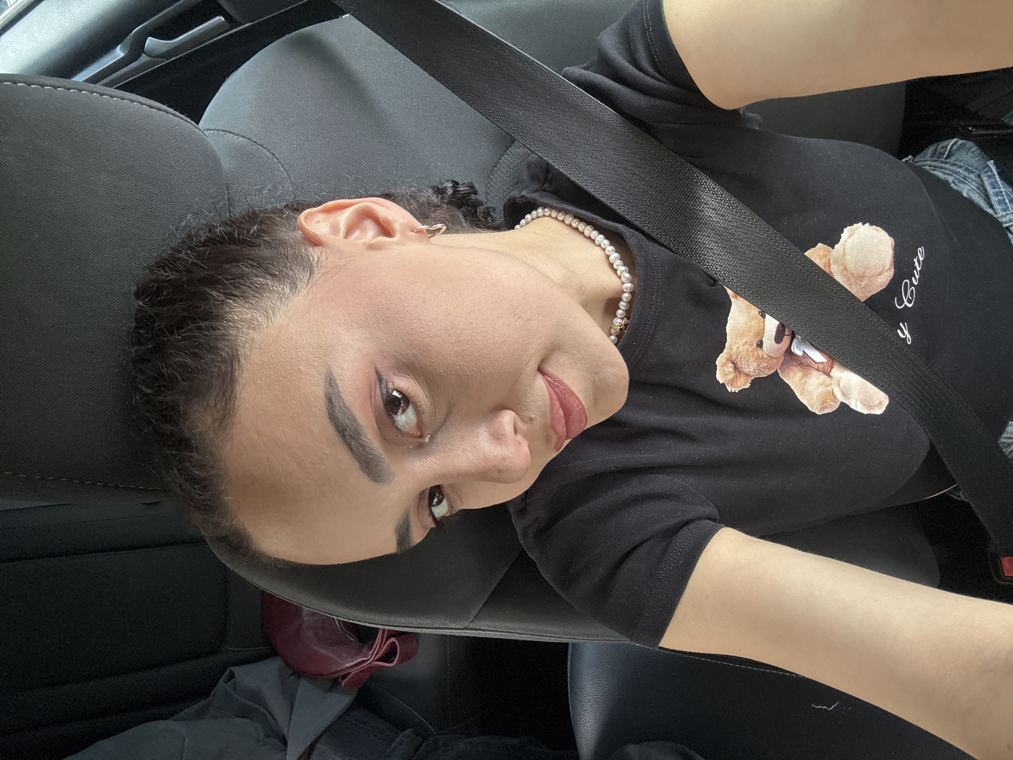 The image shows a woman inside a car wearing a black t-shirt with a teddy bear graphic. She is also wearing a pearl necklace. A seatbelt is across her body. The car seats are dark gray. There are personal items such as a red bag and clothing in the backseat. The location is not evident from the image, but the city is specified as Tbilisi, Georgia. There is no concrete information that indicates the image was taken there.