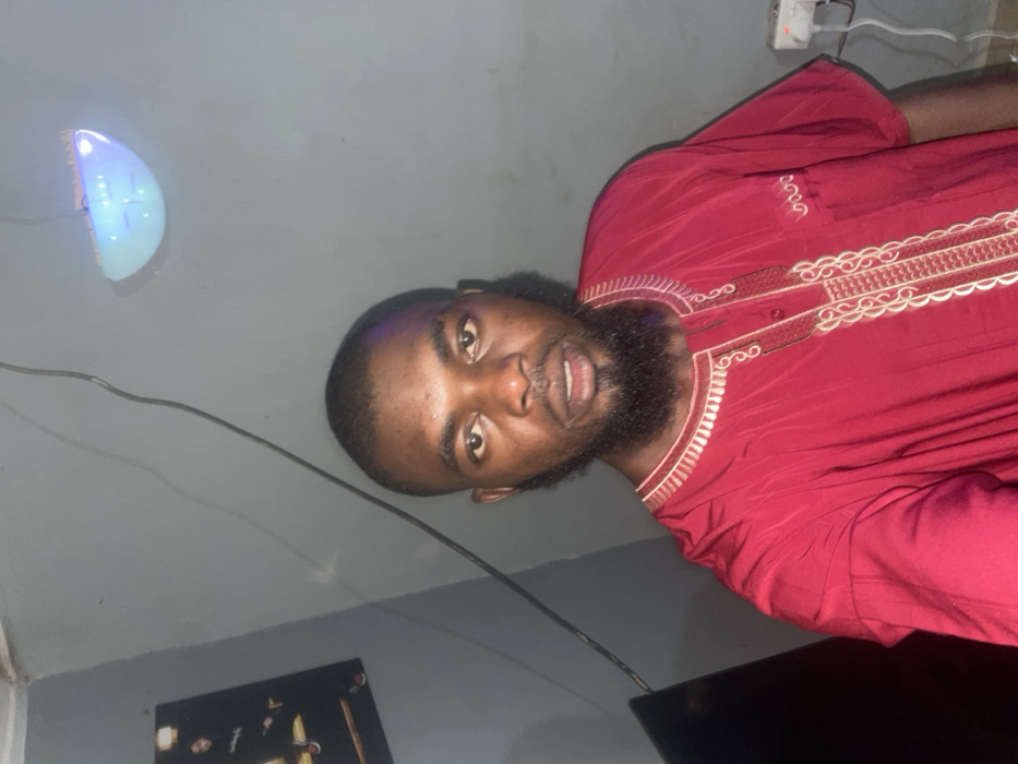 A bearded man in a maroon shirt with gold embroidery is posing for a photo. The photo is rotated counter-clockwise. He has dark skin and short hair. Behind him, the gray wall features a blue light fixture and a black framed picture. A white power strip is visible near the top corner. The overall lighting is low, casting shadows on the wall.