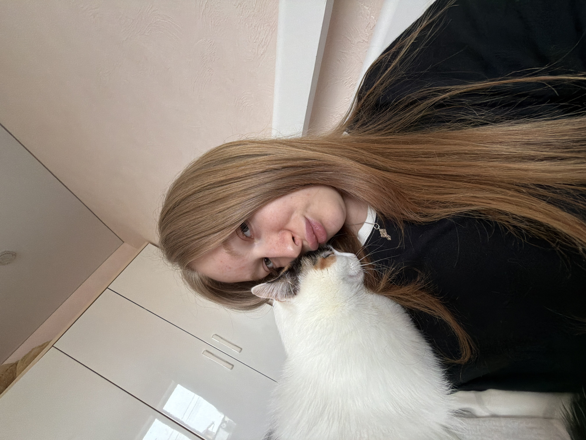 A woman is seen at a close angle, with a white cat nestled near her face. She has long, straight, light brown hair and is wearing a black top. A white cabinet with closed doors is in the background, situated adjacent to a white ceiling. Based on the available information, a location in the city of Rossiyskiy, Russia, cannot be ascertained.
