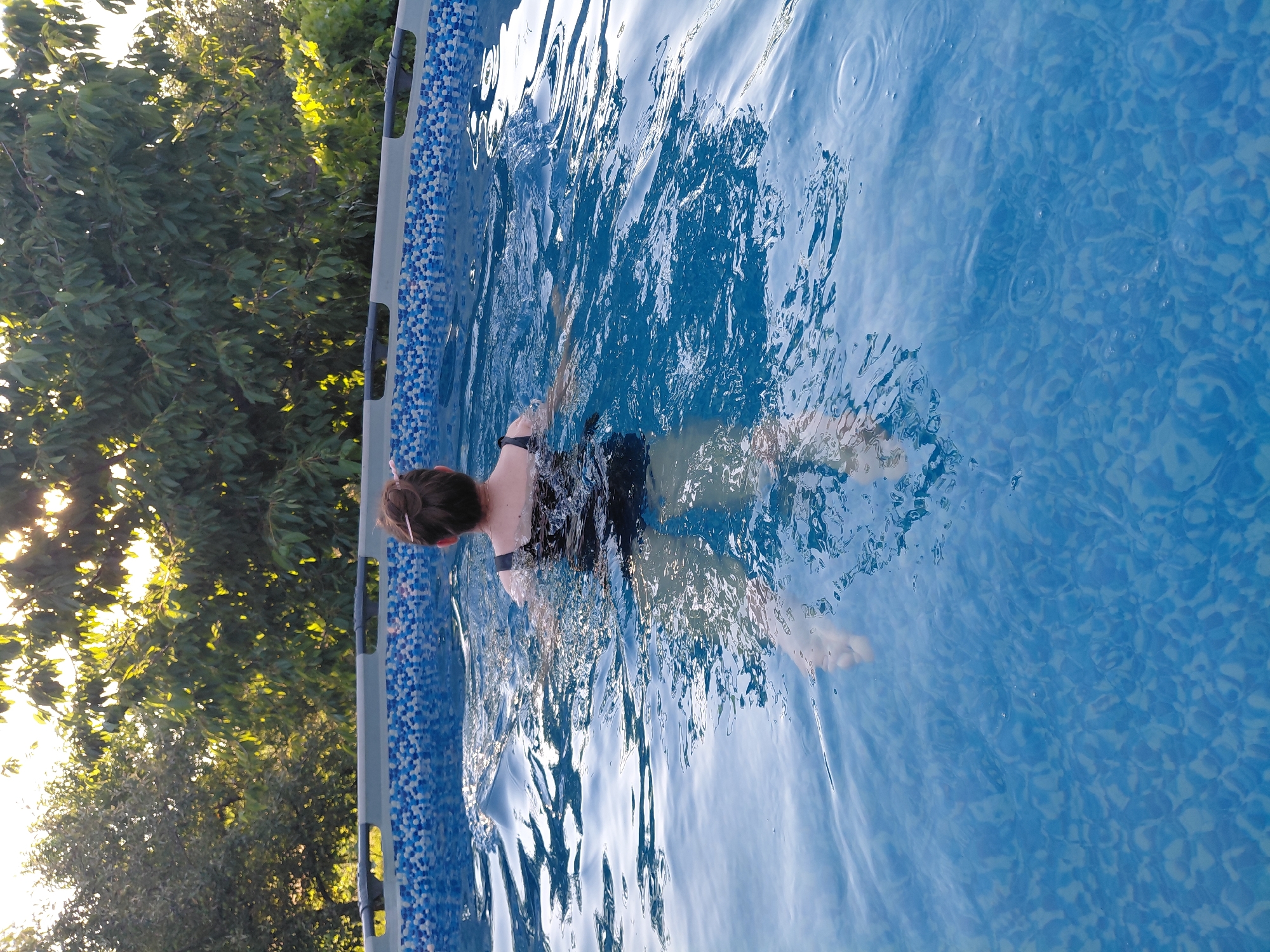A person swims in a blue, above-ground pool. The person is wearing a black swimsuit. The water is clear and blue, with sunlight reflecting off the surface, creating ripples and patterns. The pool has a blue and white tiled pattern along the top edge. To the left of the pool, there are lush green trees with sunlight filtering through the leaves, suggesting it is daytime. The overall scene appears to be outdoors in a backyard or garden setting, evoking a sense of relaxation and summer fun. The person's posture suggests they are actively swimming, possibly a freestyle stroke given the arm movement visible. The lighting indicates it is likely late afternoon or early evening, as the sun is low in the sky. No text is visible in the image.