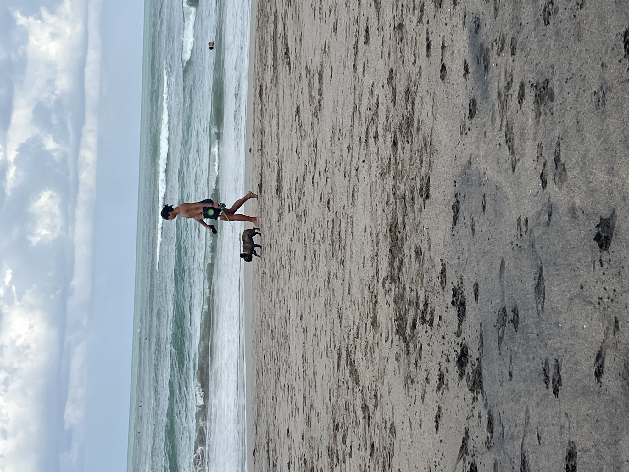 In North Kuta, Indonesia, a person walks a small dog on a sandy beach. The person, wearing dark swim trunks with a green accent, is walking from left to right. A small, dark-colored dog is on a leash, walking beside and slightly ahead of the person. In the background, the ocean waves are visible, with several surfers in the distance. The sky is overcast with clouds. The sand in the foreground shows various footprints and patches of wet sand.