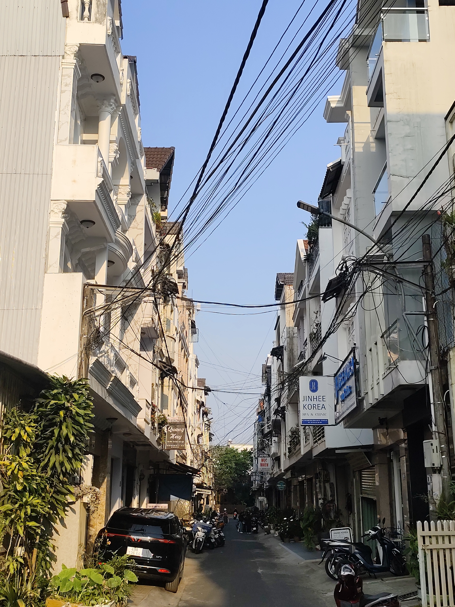 The image depicts a narrow street in Da Lat, Vietnam, lined with buildings of varying heights and architectural styles. The buildings are predominantly white or light-colored, with some exhibiting decorative balconies and ornate details. A dense network of overhead electrical wires crisscrosses the sky above the street.
The street is paved and appears to be used by both pedestrians and vehicles. Several motorcycles are parked along the sides of the street, and a black SUV is parked on the left. Signage for businesses, including "JINHEE KOREA SPA & CLINIC" and "QUANG HUY" are visible on the buildings. Green foliage is present on the left side of the street, adding some natural elements to the urban setting. The sky is a clear, light blue, suggesting a sunny day. The scene is typical of the urban landscape in Da Lat, with a mix of commercial and residential buildings, dense infrastructure, and a busy street life.