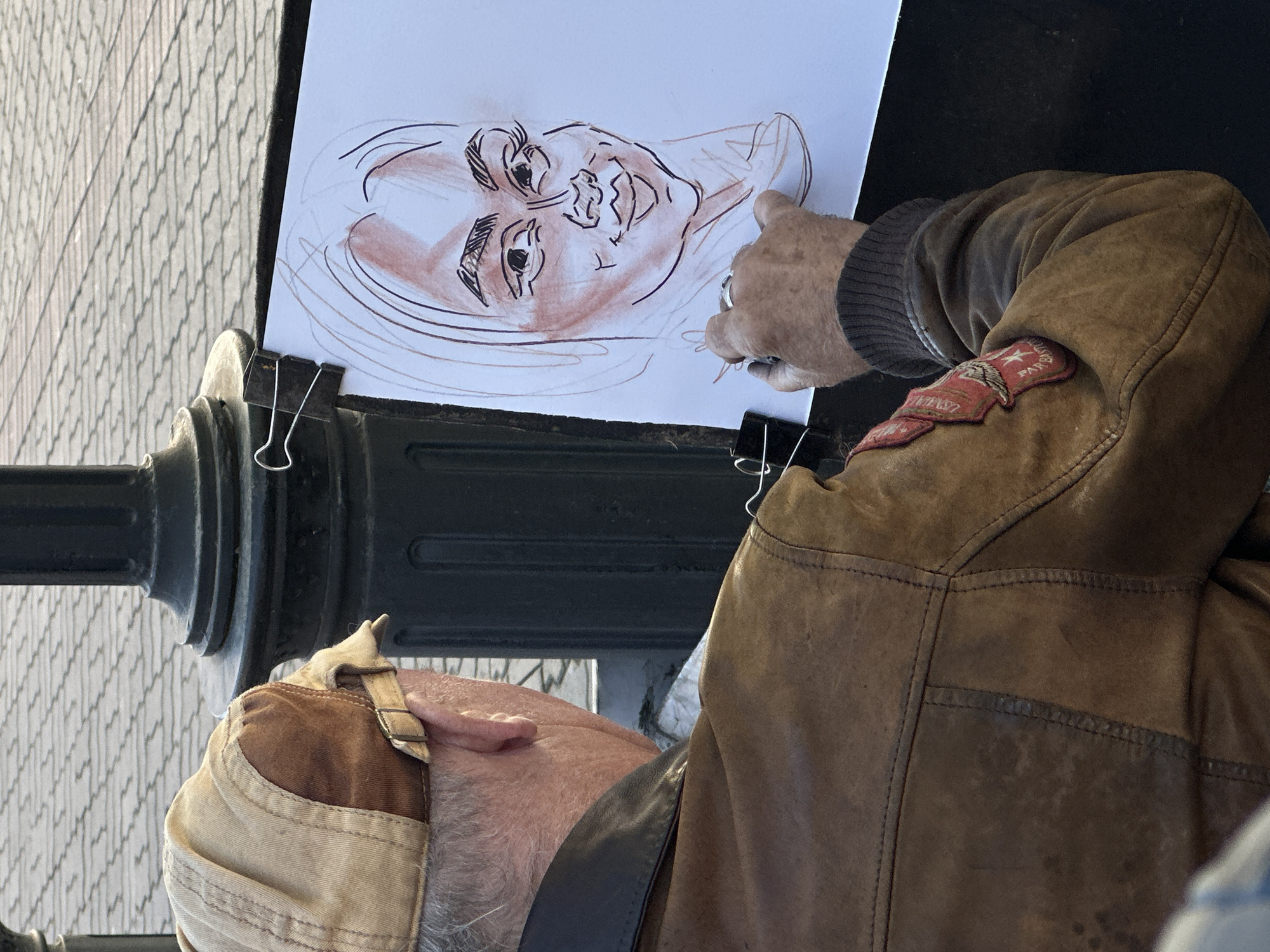 A man, positioned from the shoulder up and facing right, is drawing a caricature on a sheet of white paper clipped to a dark, possibly metallic, structure. The drawing, executed in shades of brown and black, features a face with exaggerated features. The man's right hand, adorned with a ring, holds a drawing tool. He wears a brown leather jacket with a red patch on the right shoulder bearing a star and possibly some text. A cap is visible on his head. The setting appears to be outdoors, as suggested by the brick-like texture in the background. The location is Kazan, Russia.