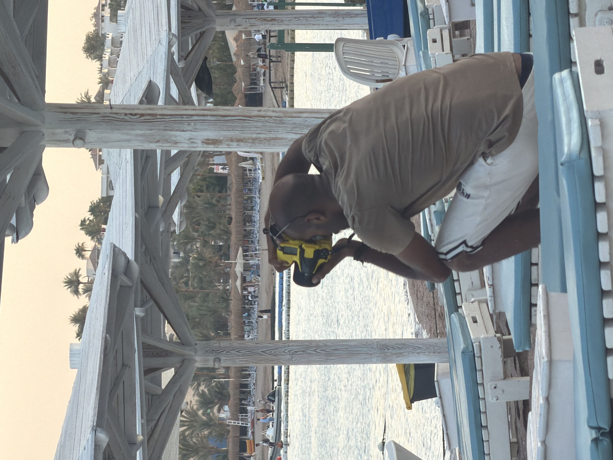 A man is kneeling on a beach in Sharm El Sheikh, Egypt, holding a yellow camera to his eye and taking a picture. He is wearing a brown t-shirt and white shorts. In the background, there is a beach with rows of lounge chairs and umbrellas, palm trees, and buildings. The sea is visible, with boats on the water. The sky is overcast with a light hue.