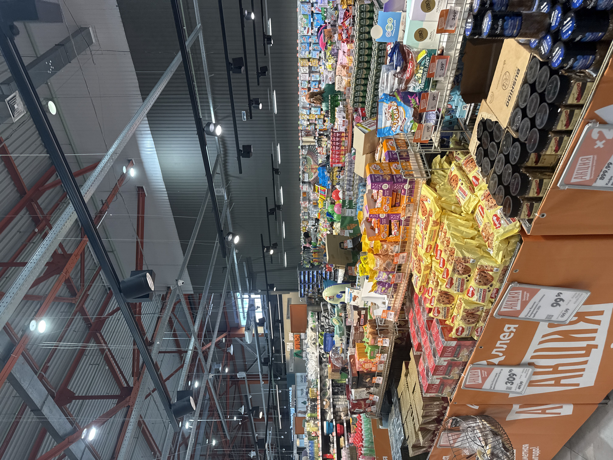 This image shows the interior of a large supermarket or hypermarket, likely in Angarsk, Russia, given the Cyrillic text. The perspective is from a high vantage point, looking down the aisles, and the image is rotated 90 degrees clockwise.
The aisles are stocked with a wide variety of consumer goods, including packaged foods like cookies, candy, and snacks, as well as beverages and other household items. There are promotional signs and displays throughout the store, indicated by the prominent "АКЦИЯ" (Aktsiya), meaning "Sale" or "Promotion" in Russian. Some of these signs feature prices like "99", "109,99", "179", "259", "309", "349", "499,99", and "899,99". Brand names visible include "LEBO", "Jardin", "BUSHIDO", "TESS", "КОРОВКА" (Korovka - a type of candy), and "Победа" (Pobeda - Victory).
The lighting appears to be artificial, typical of an indoor retail environment, with rows of spotlights visible on the ceiling structure. The architecture suggests a modern, expansive building with exposed metal beams and a corrugated metal ceiling.
There are no people clearly visible in the frame, but the presence of merchandise and promotional materials indicates that the store is open and likely has customers browsing, even if they are not in focus or easily discernible from this angle. The overall impression is of a busy commercial space stocked for shoppers.