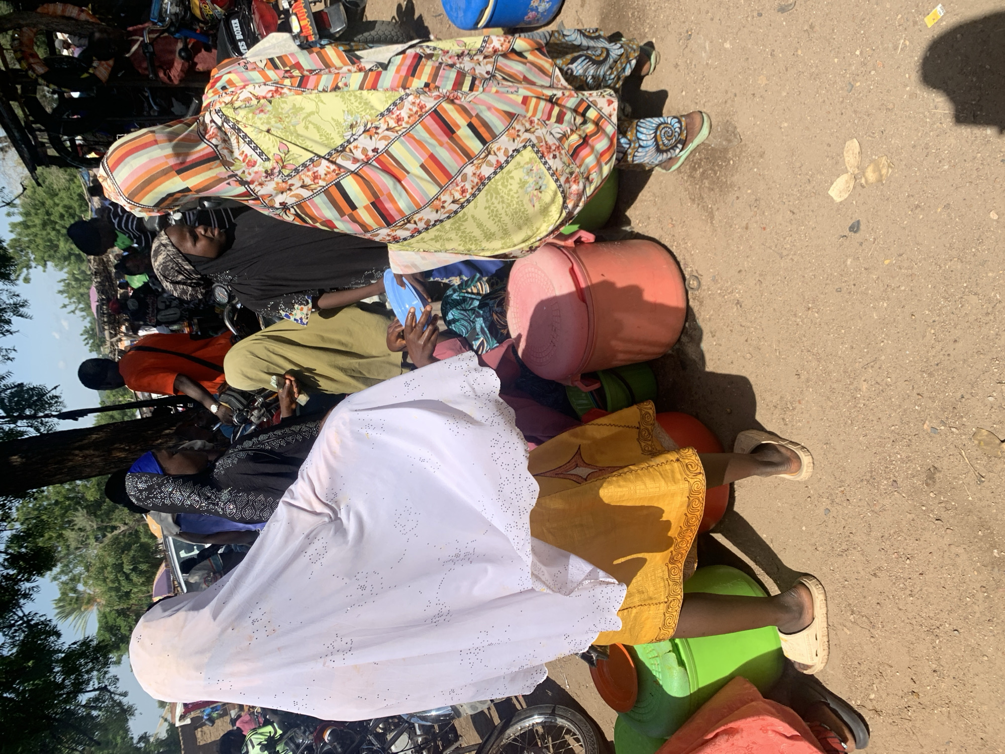The image depicts a group of people, likely in Boi A, Nigeria, gathered around what appears to be a transportation hub or marketplace. The orientation of the image is vertical, with the subjects appearing in a somewhat jumbled and layered arrangement.
The main subjects are several individuals, primarily women, dressed in colorful and patterned clothing, including headscarves and long dresses. One woman in the foreground is wearing a white garment adorned with small, dark decorative elements. Another is wearing a vibrant, patchwork-style dress. A woman in a black garment and headscarf is visible further back.
Numerous objects are scattered amongst the people, including large plastic containers in various colors (blue, green, pink, and orange), likely used for storage or transport. Some of these containers are quite large and seem to be serving as makeshift seating. There are also what appear to be bags and possibly parts of motorbikes, suggesting this is an area where people are preparing to travel or have just arrived. One person is holding a small blue object.
The setting appears to be an outdoor, unpaved area, with sand and dirt visible on the ground. Sunlight casts distinct shadows, indicating it is daytime, likely mid-morning or afternoon given the intensity of the shadows. The presence of trees and foliage in the background suggests a rural or semi-rural location.
The activity seems to be related to travel or waiting for transportation, with people sitting on or near their belongings. The overall mood is difficult to ascertain due to the angle and cropping, but the scene appears somewhat crowded and informal. There are no discernible emotions expressed by the individuals. No visible text is present in the image.