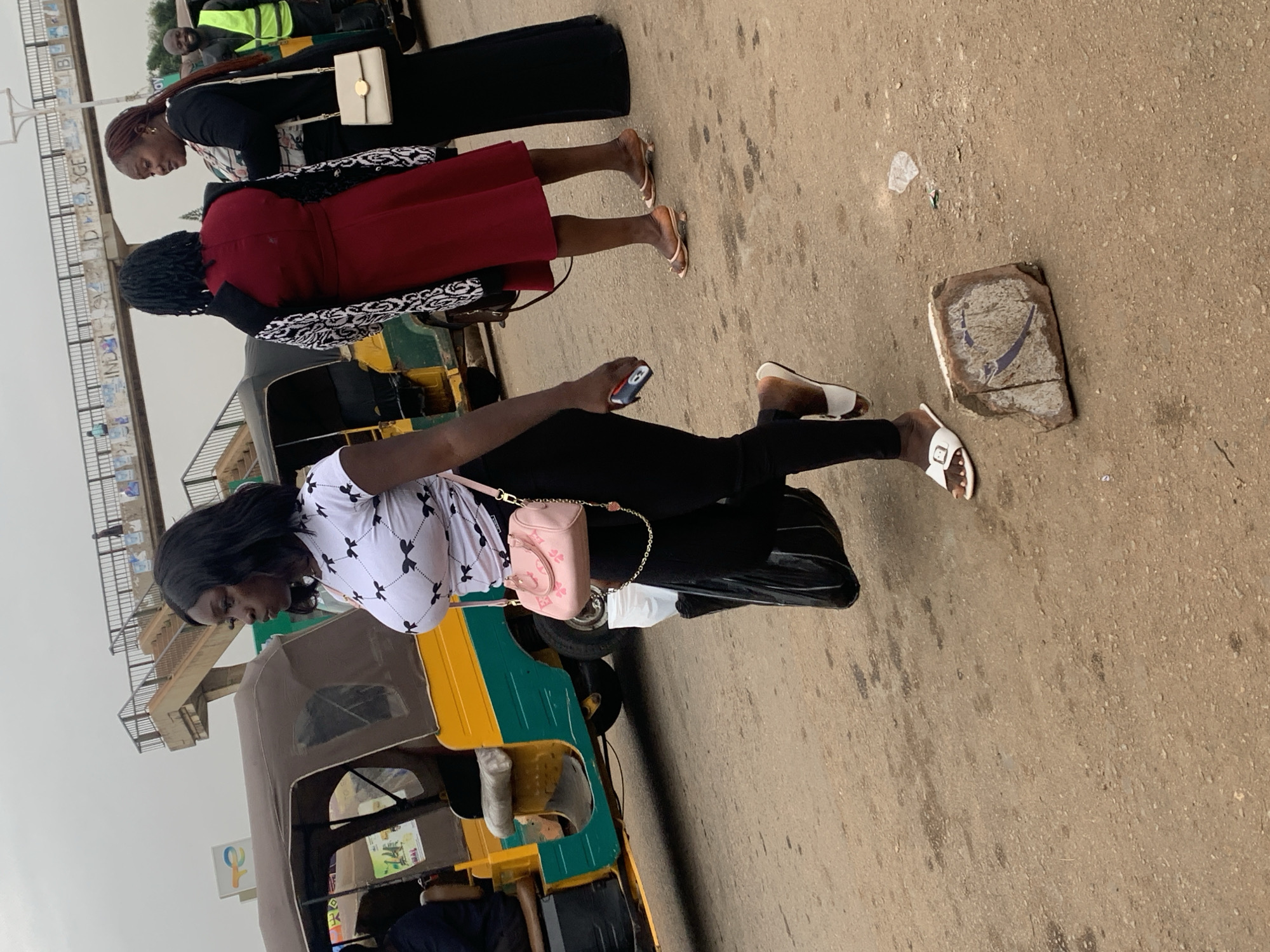 In Gura Top, Nigeria, a woman with dark skin and black hair walks along a dirt road. She is wearing a white t-shirt with a black bow pattern and black leggings, along with white sandals. She carries a black plastic bag in her left hand and a pink purse with a gold chain on her right shoulder. In the background, other people are visible, including a woman in a red dress and a woman in a black dress. A three-wheeled vehicle, possibly a taxi, is parked on the left side of the road. An elevated walkway or bridge with stairs is visible further in the background, along with some graffiti. The ground is dusty and uneven, with some debris scattered around.